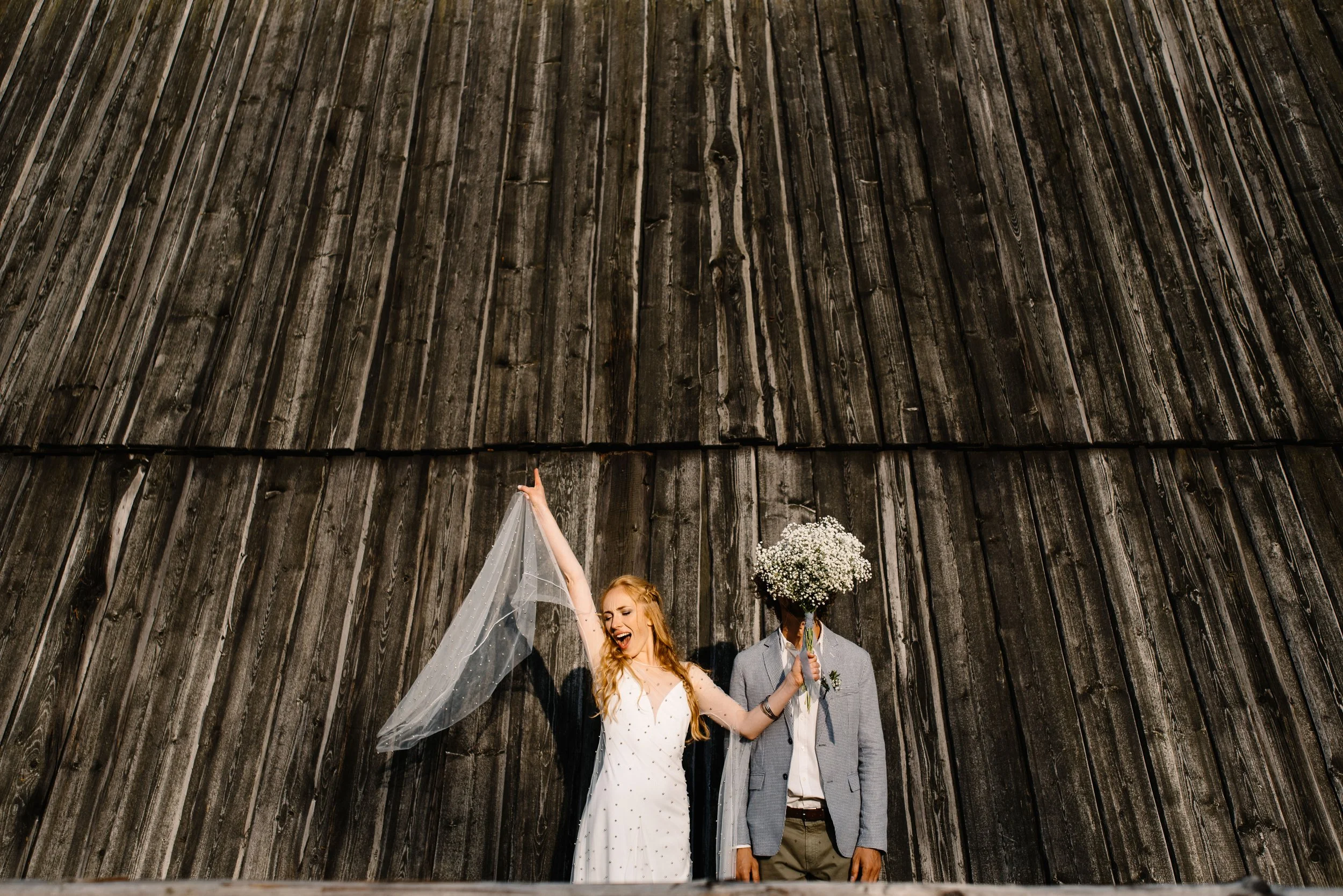 A newlywed couple standing in front of a large wooden wall. The bride is smiling, wearing a white wedding dress and holding a sheer veil, raising her hand. The groom, whose face is hidden by a bouquet of white flowers, wears a gray blazer and white shirt.