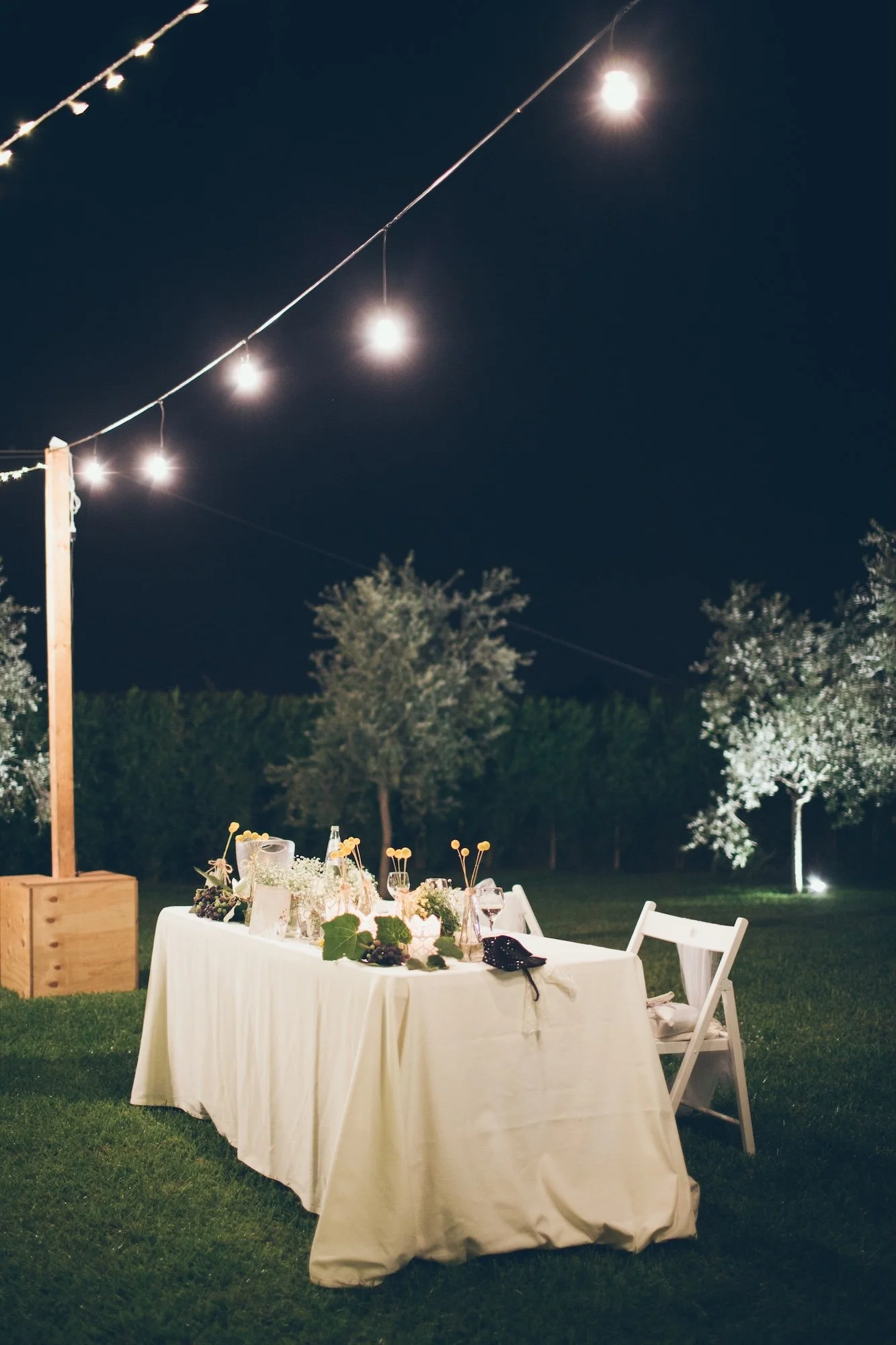 An outdoor dinner table set under string lights at night, with a white tablecloth, chairs, and decorative items, surrounded by trees and greenery.