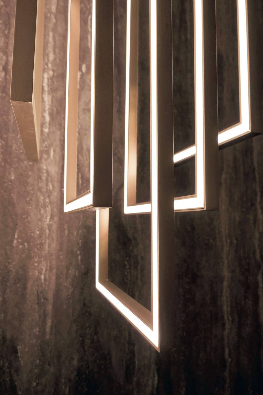 Modern geometric vertical brass installation with illuminated rectangular frames against a dark textured wall.