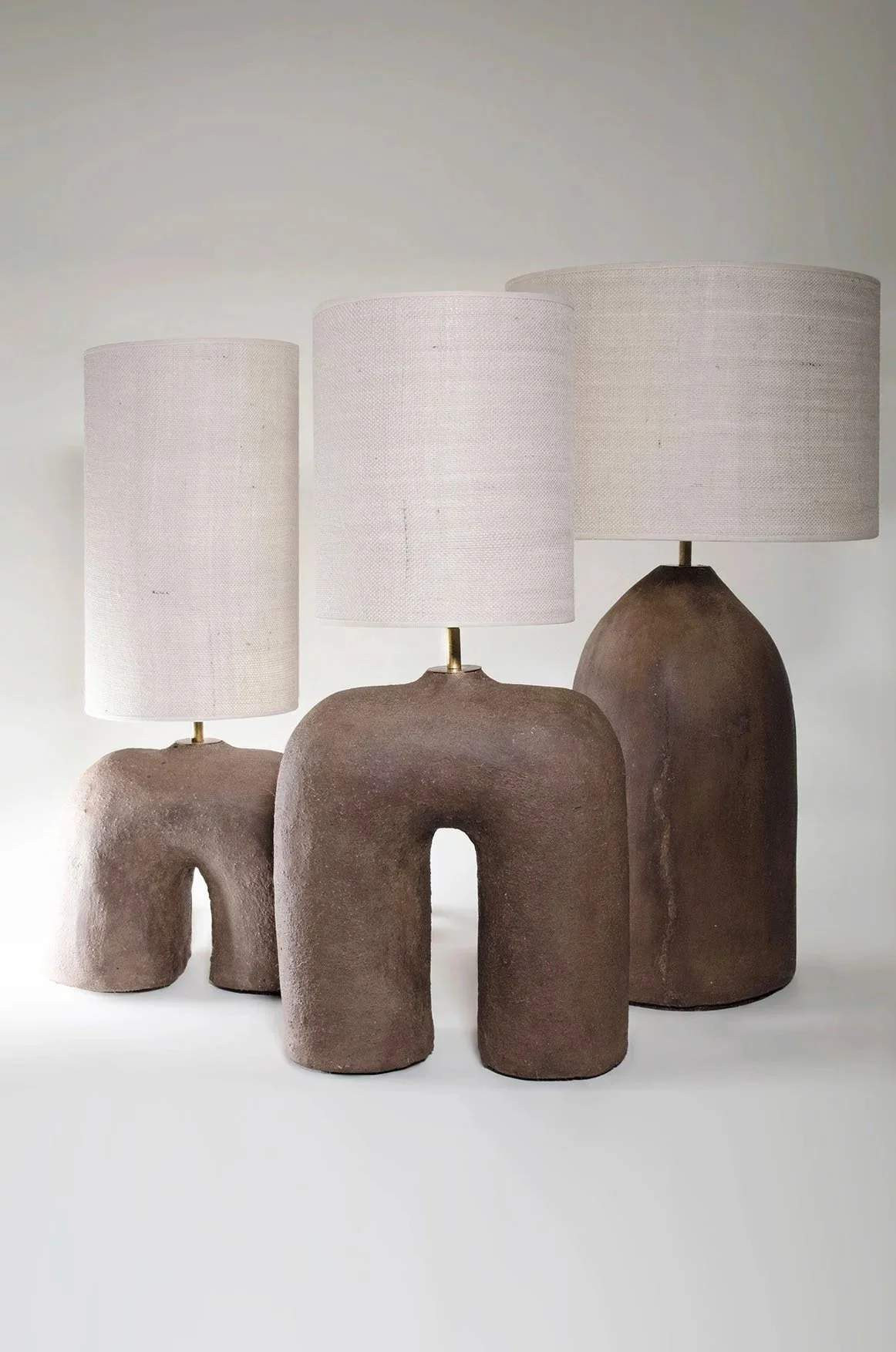 Three Philokalia table lamps by Studio Vourli, featuring beige fabric shades and hand-formed cob bases in three distinct sculptural shapes against a neutral background.