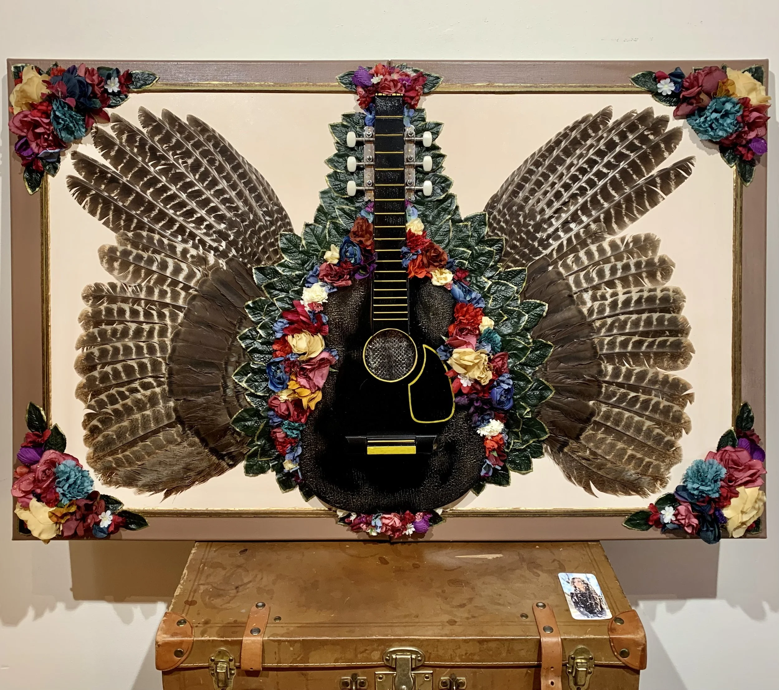 Artwork featuring a black guitar adorned with colorful flowers and large brown bird feathers against a beige background, framed with floral decorations, resting on a vintage suitcase.