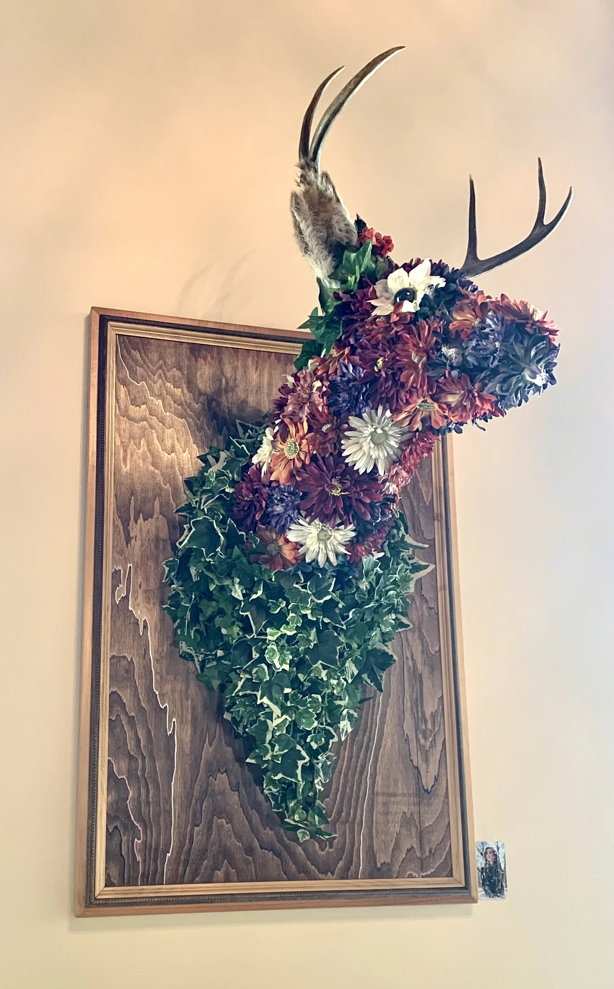 A deer head sculpture mounted on a wooden plaque is decorated with flowers and leaves. The antlers are natural, but the head and neck are covered in colorful artificial flowers and green foliage, creating a nature-themed artistic display on the wall.