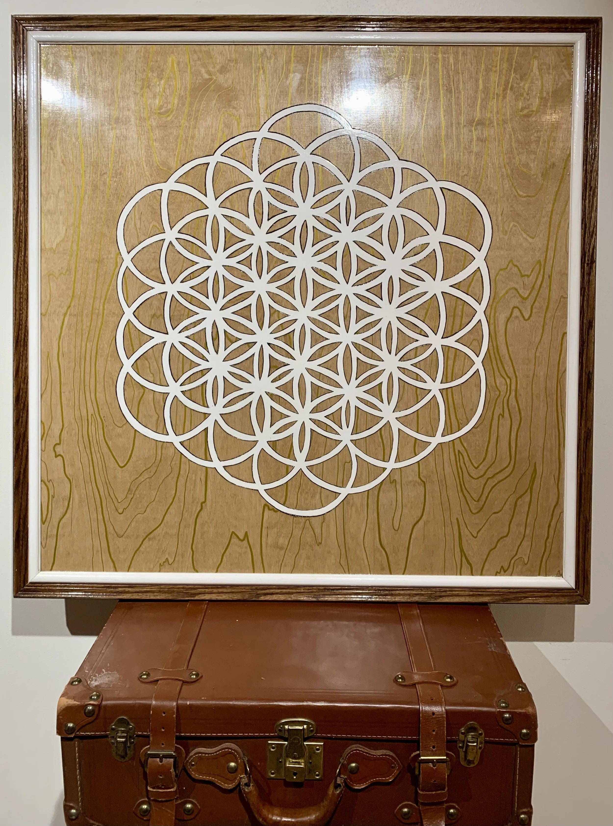 Abstract geometric pattern resembling the Flower of Life in a white outline on a wood-textured background, framed and displayed above a leather suitcase.