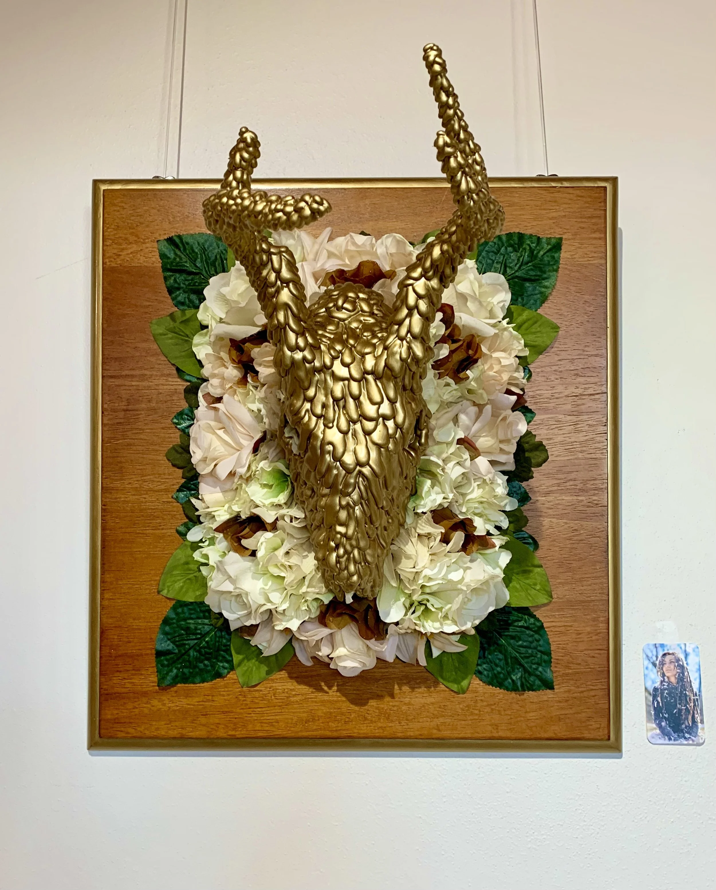 Wall-mounted art piece featuring a gold animal skull with antlers, surrounded by white flowers and green leaves, set against a wooden background.