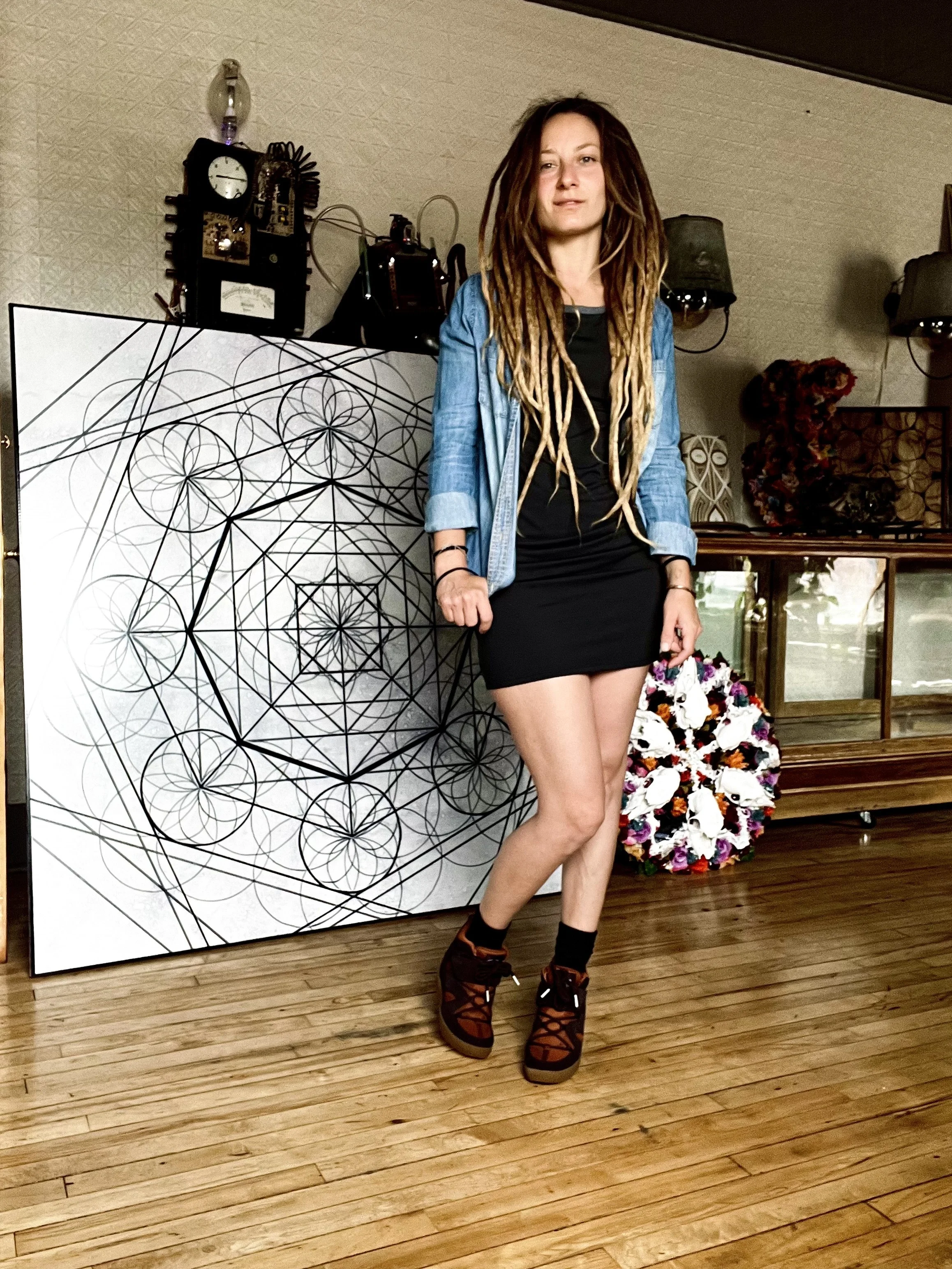 Woman with long hair standing indoors next to a geometric art piece, holding a colorful circular object, with vintage decorations behind.