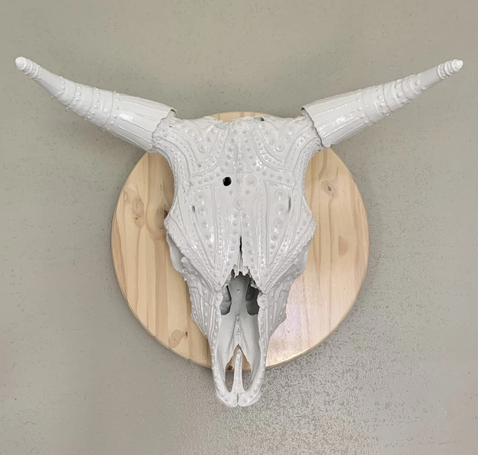 Decorative white bull skull with engraved patterns mounted on a wooden plaque, displayed on a wall.