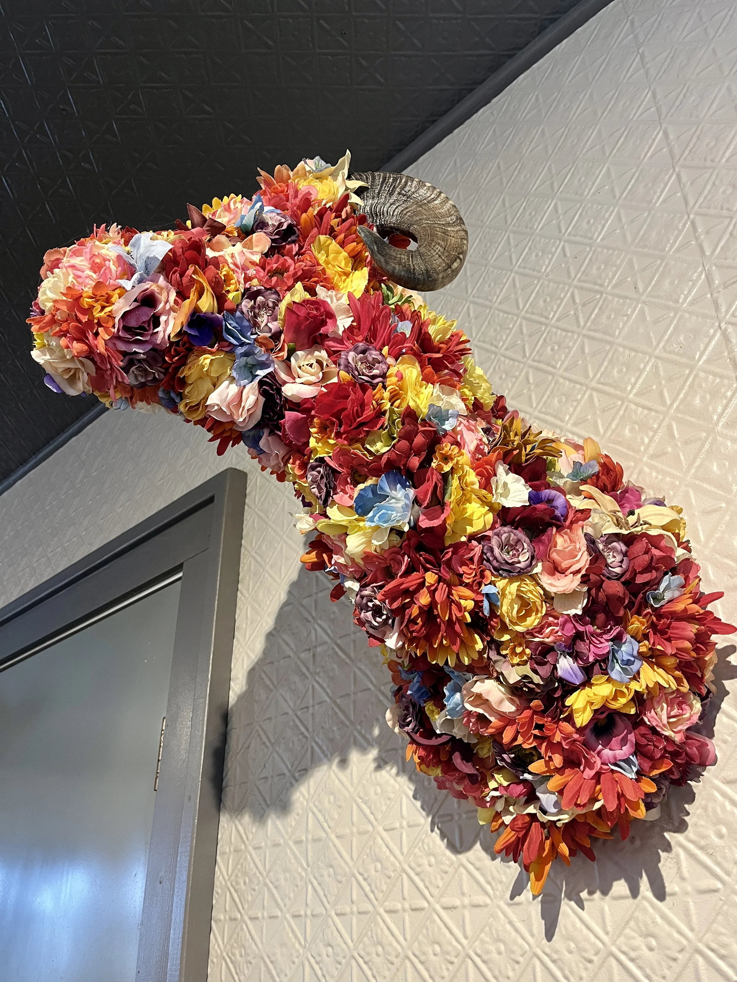 Wall-mounted ram head sculpture covered in colorful artificial flowers.