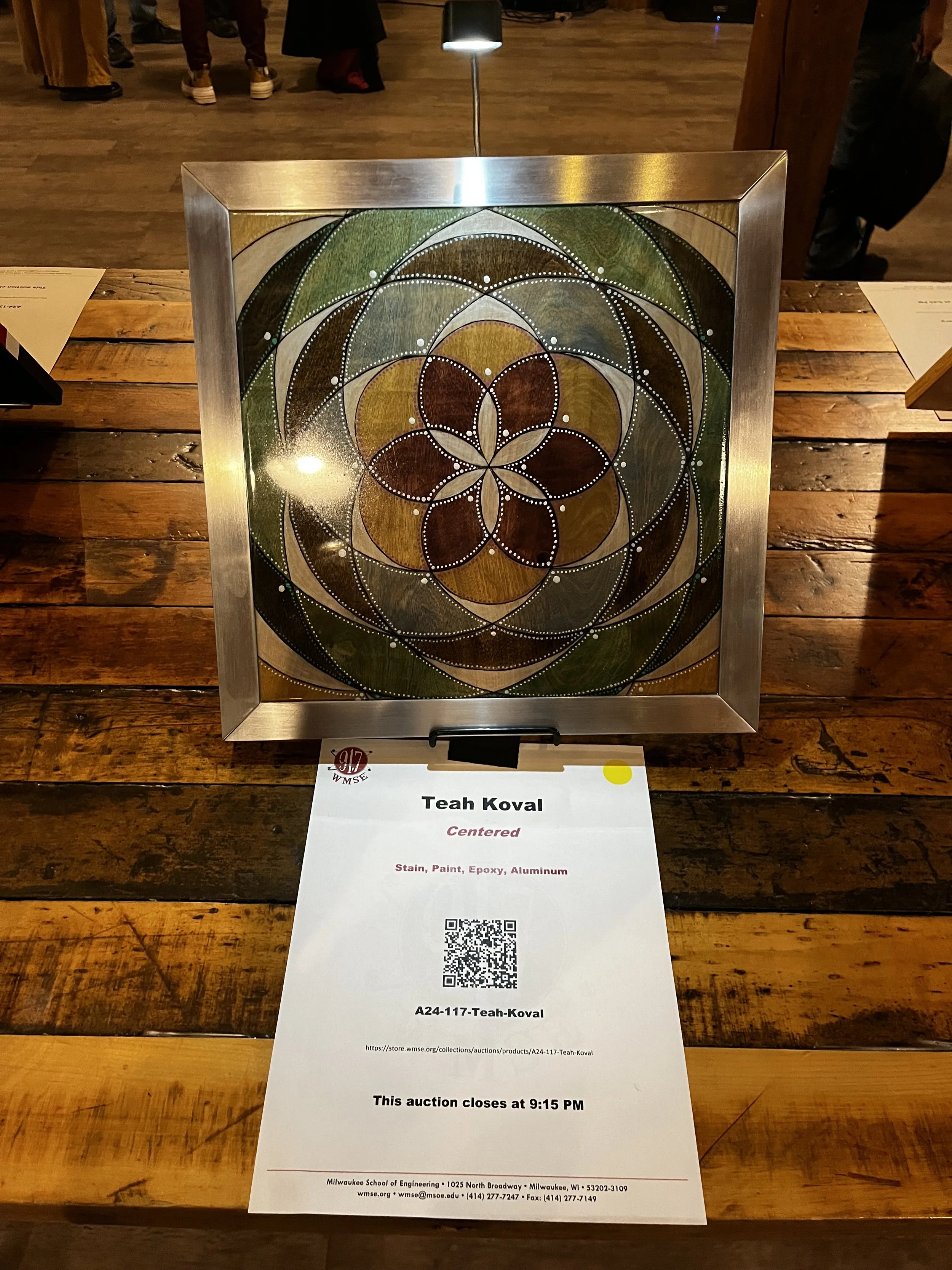 Framed geometric artwork on a wooden table, featuring a circular pattern with intersecting shapes in earth tones. Below is a paper with details about the artist, "Teah Koval," and auction information, including a QR code.