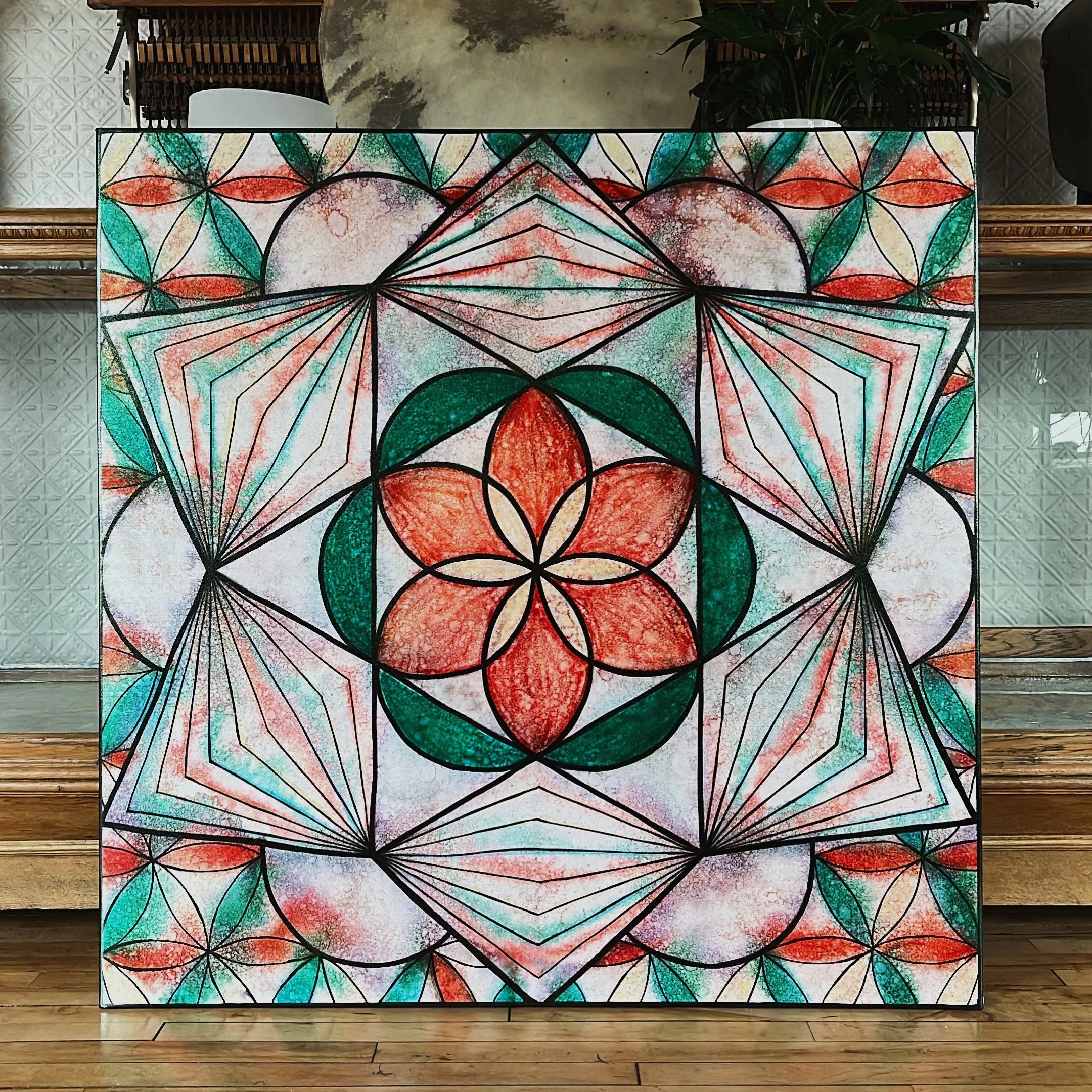 A colorful geometric and floral abstract artwork with red, green, and orange hues, featuring interlocking shapes and symmetrical patterns. The design includes a central floral motif surrounded by complex geometric shapes, creating a kaleidoscopic effect.