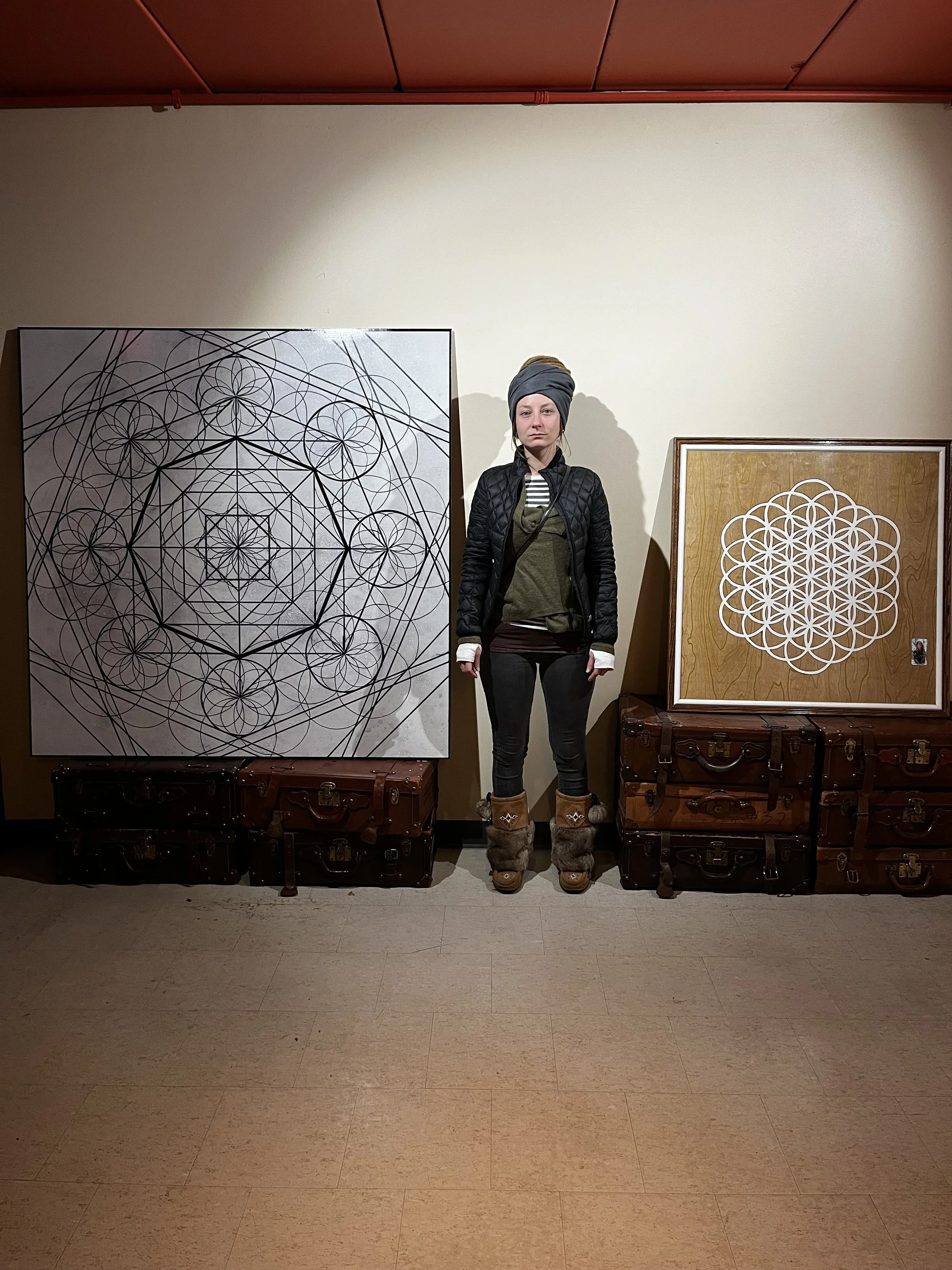A person stands in a room between two pieces of geometric artwork, with several vintage suitcases stacked beneath them. The artwork features intricate geometric patterns. The person wears a hat, jacket, leggings, and fur-trimmed boots.