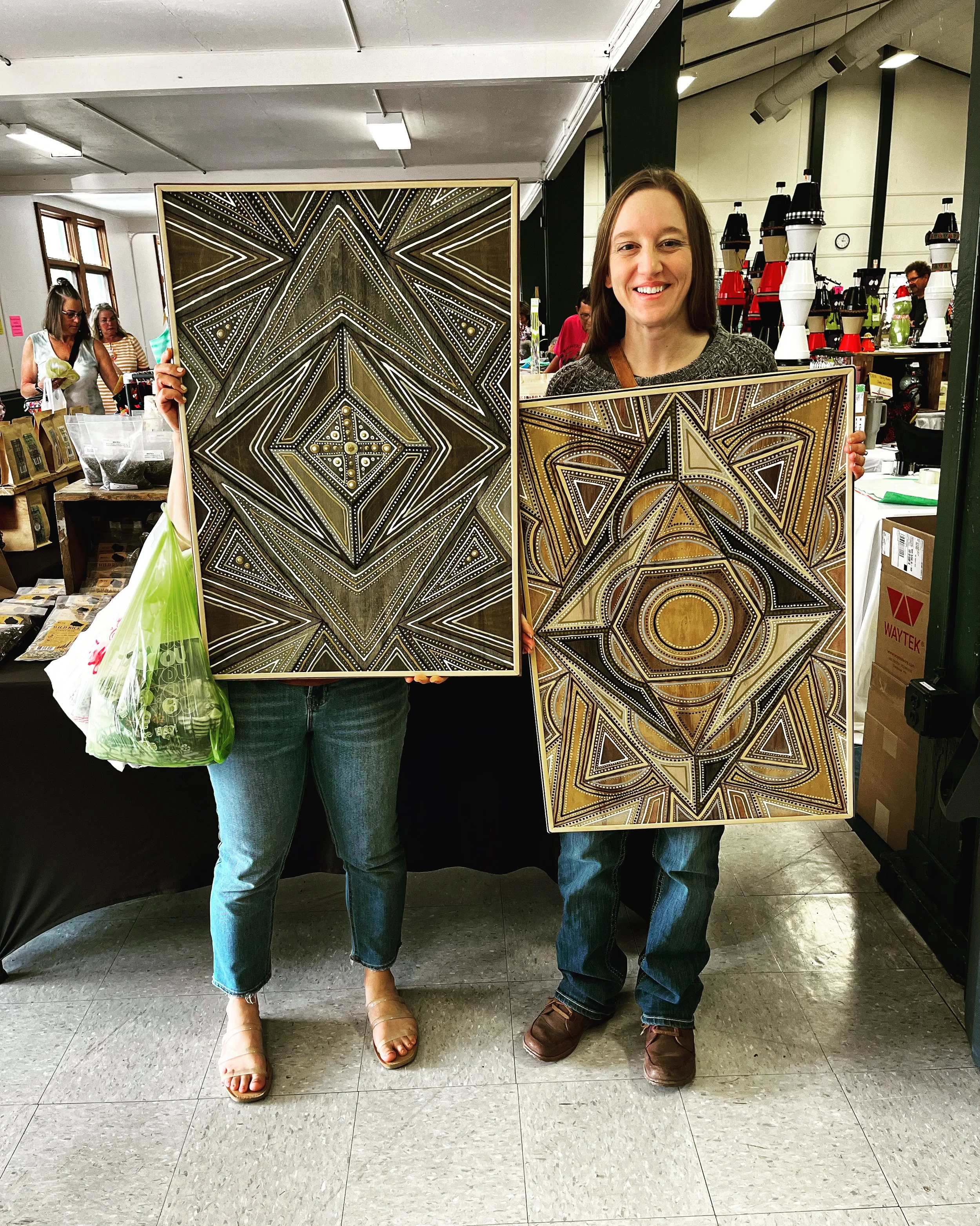 Two people holding geometric patterned artworks in an indoor market setting.