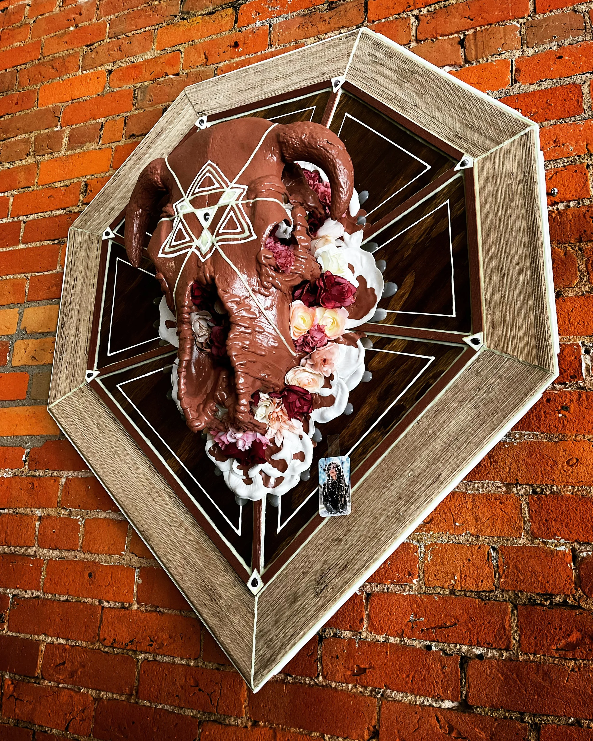 Decorative skull mounted on a brick wall, featuring a geometric design and surrounded by artificial flowers.
