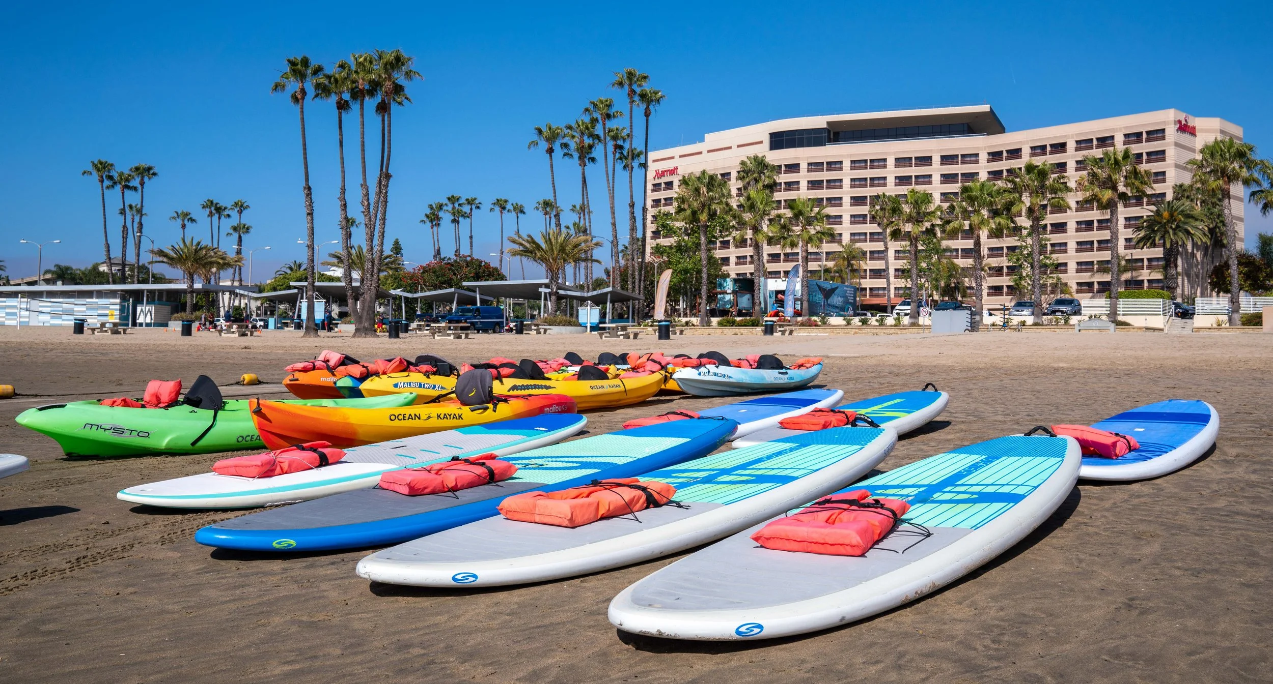 Photo by Brandon Kirk of a beautiful sunny day on Mother's Beach with vibrant set of kayaks and paddleboards in front of Pro SUP Shop with a view of the Marina del Rey Mariott