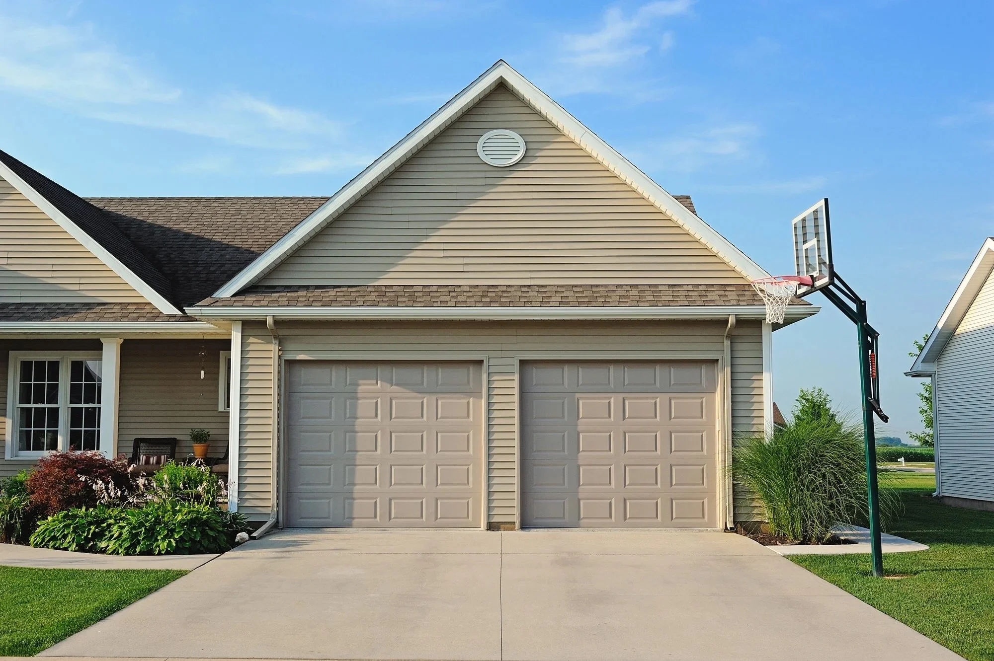 A house with double garage doors, a small porch with potted plants, and a portable basketball hoop outside on a driveway, under a clear blue sky.