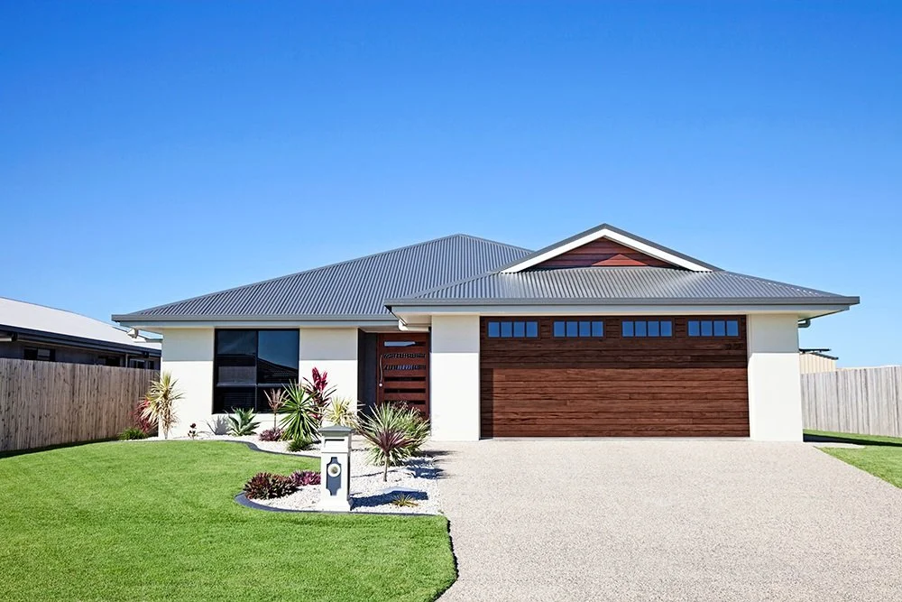 Modern house with a gray metal roof, white walls, and a wooden garage door, surrounded by a well-kept lawn and landscaped garden, under a clear blue sky.