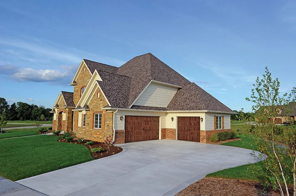 Large house with brick and siding exterior, three-car garage, and a well-maintained lawn under a blue sky.
