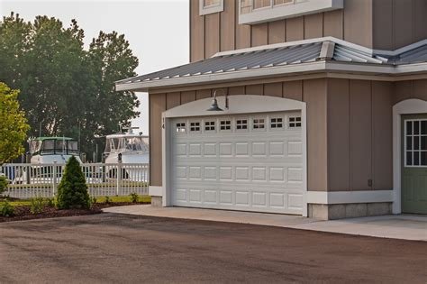A white garage door attached to a brown house with a light fixture above, situated in a residential area near boats docked by a fence.