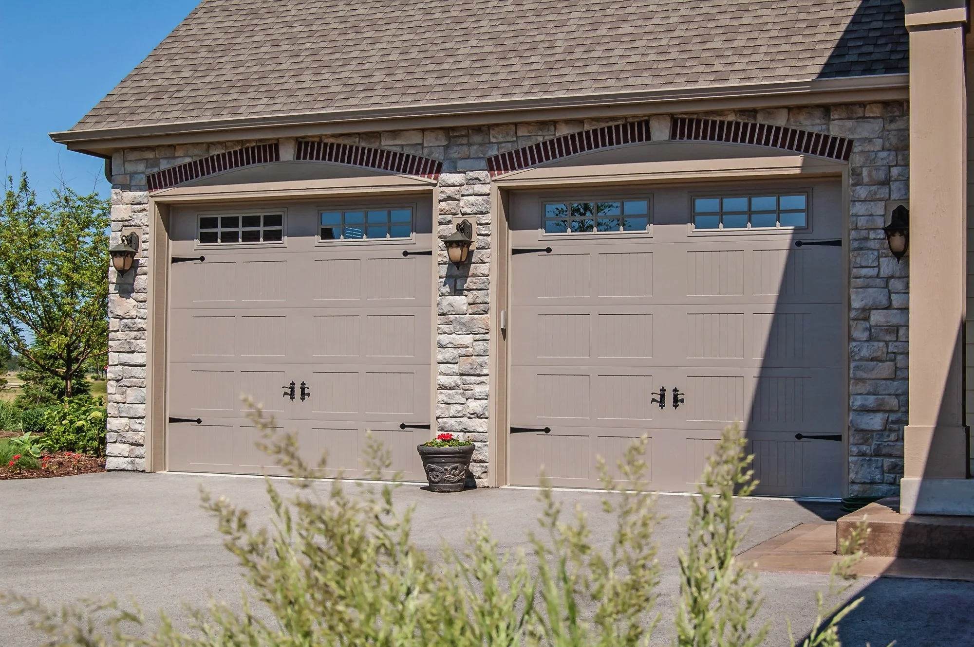 Two beige garage doors with small windows on top, mounted on a stone house. The house has a sloped shingle roof and outdoor wall lanterns. In front, there are plants and a small flower pot.
