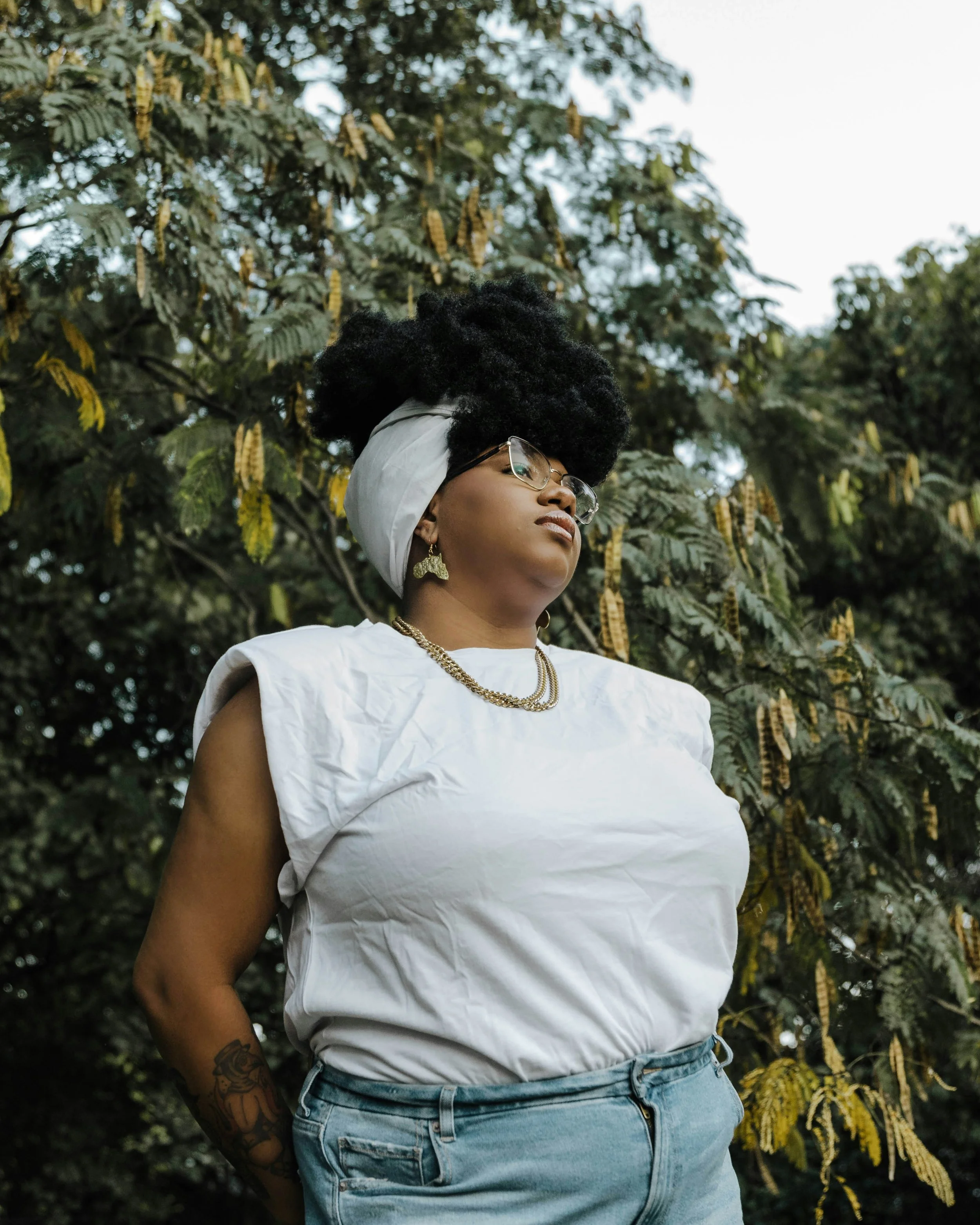 A Black person in a white shirt stands in front of a tree
