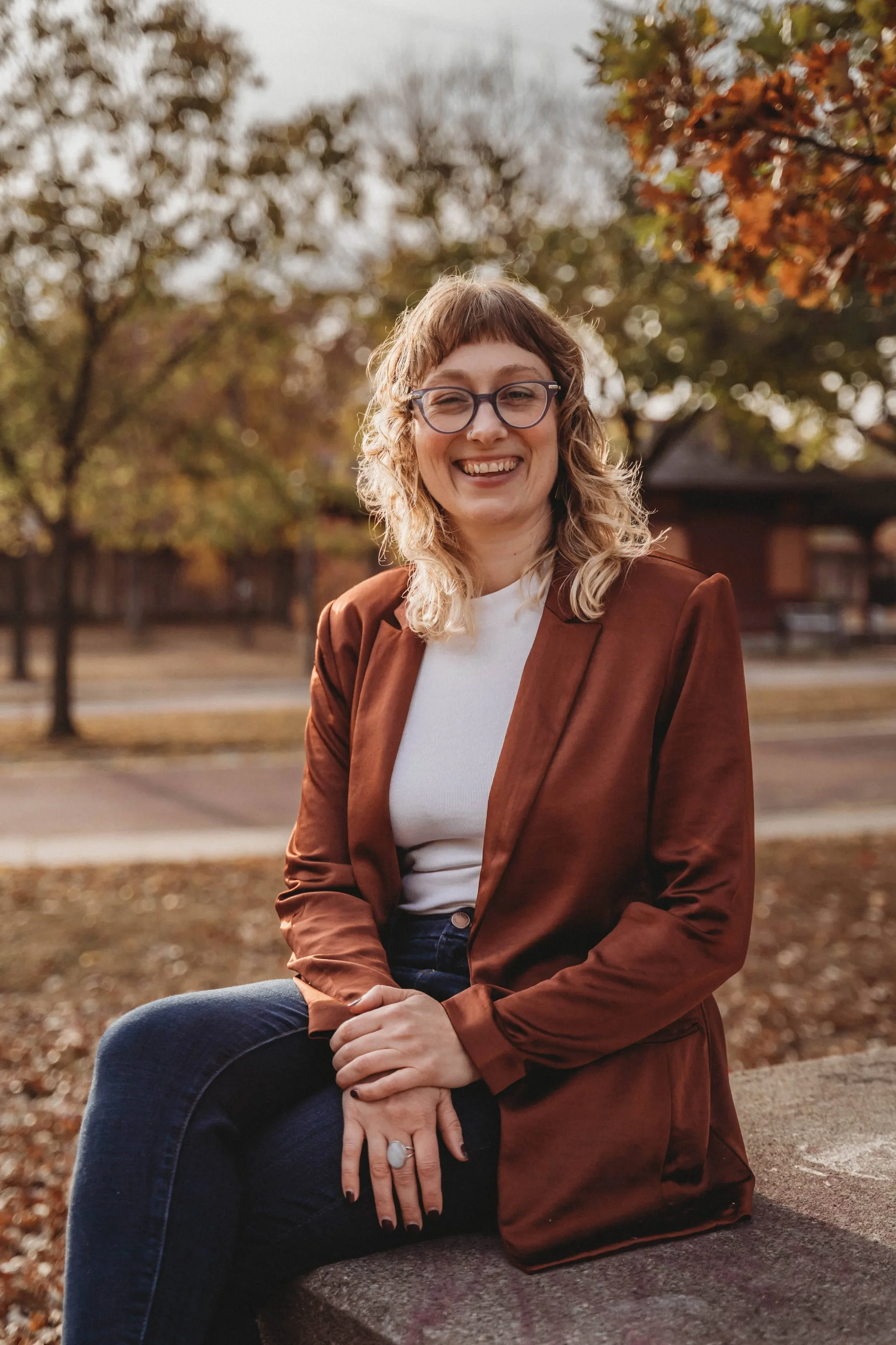 Sarah sits outside and smiles in a brown blazer (trauma therapy minneapolis)