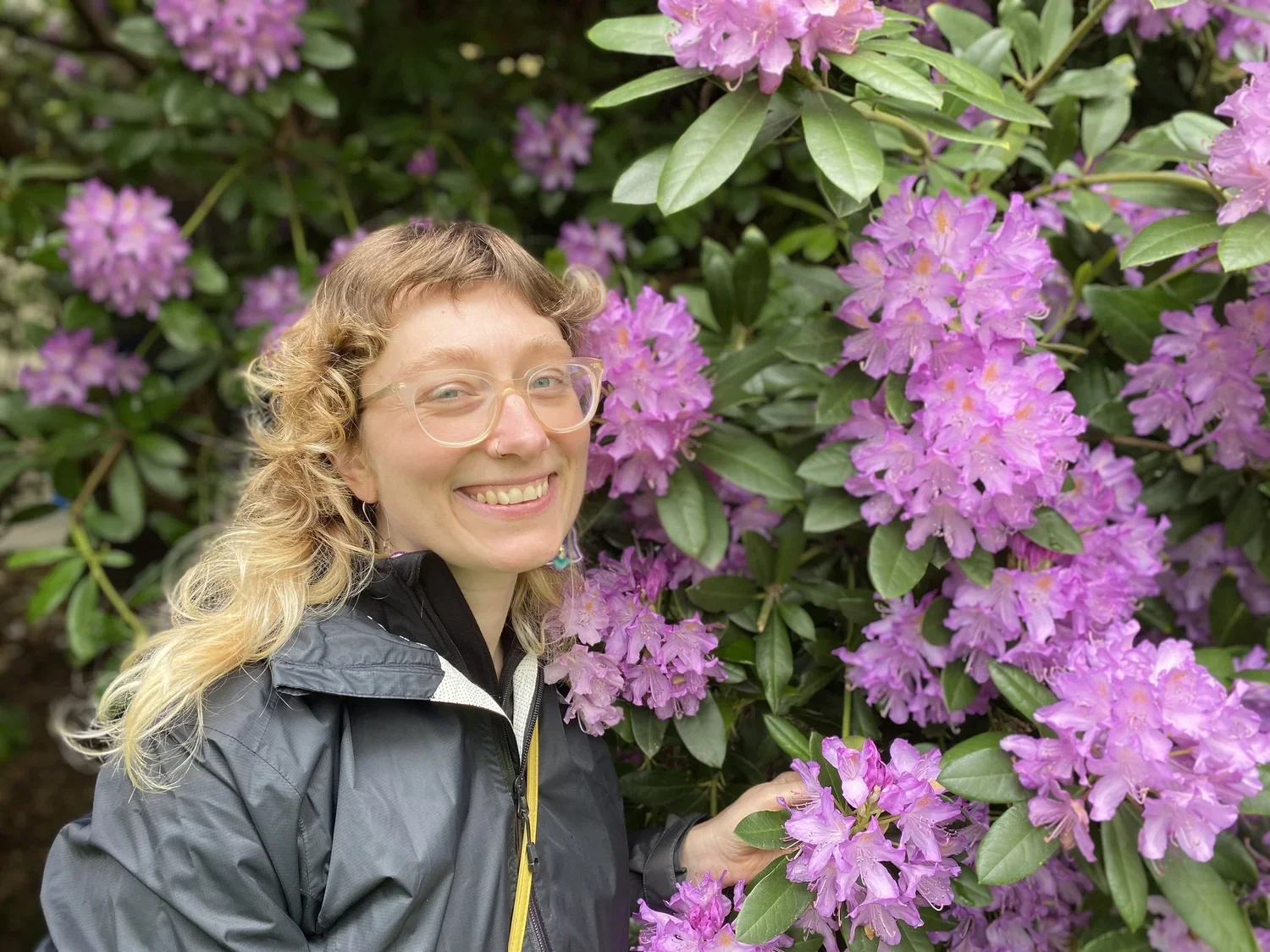 Sarah Lazarewicz, a trauma specialist and somatic therapist in St. Paul MN smiles next to a wall of purple flowers and green leaves