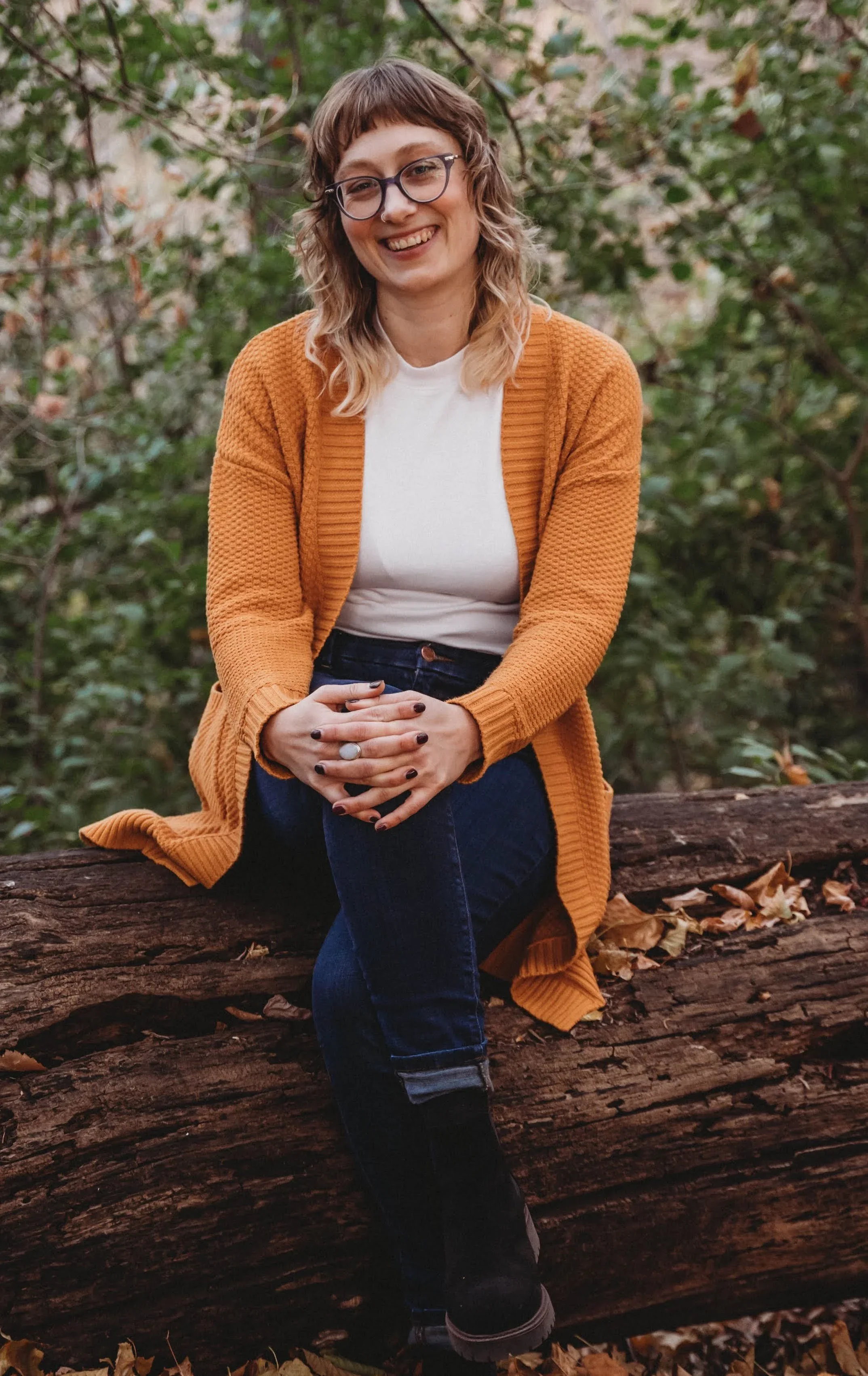 Sarah sits on a log, smiling in a yellow cardigan
