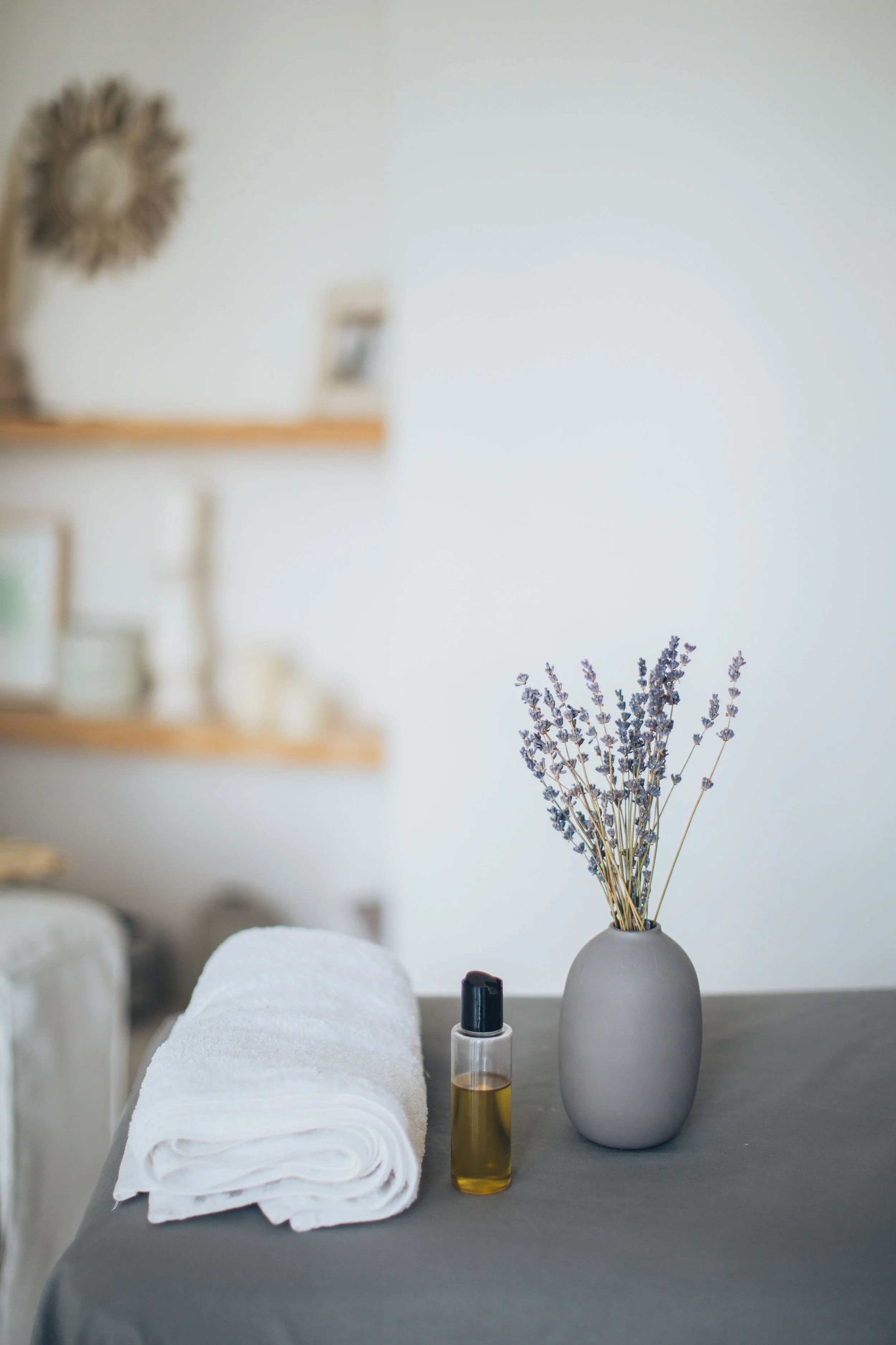 A table with a rolled white towel, a small bottle of essential oil, a spray bottle, and a vase with purple flowers. The background appears to be a living space with shelving and wall decorations.