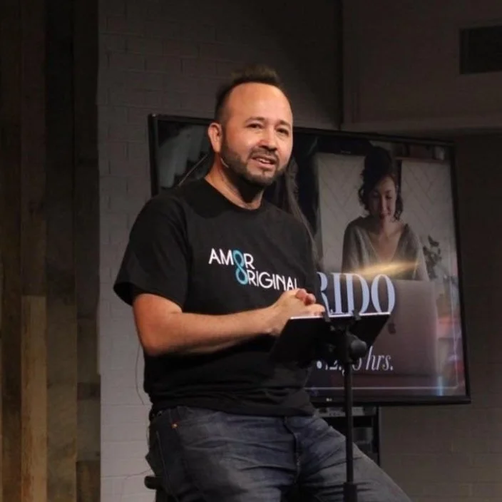 Man speaking at an event wearing 'Amor Original' t-shirt, with a microphone in front and a screen in the background.