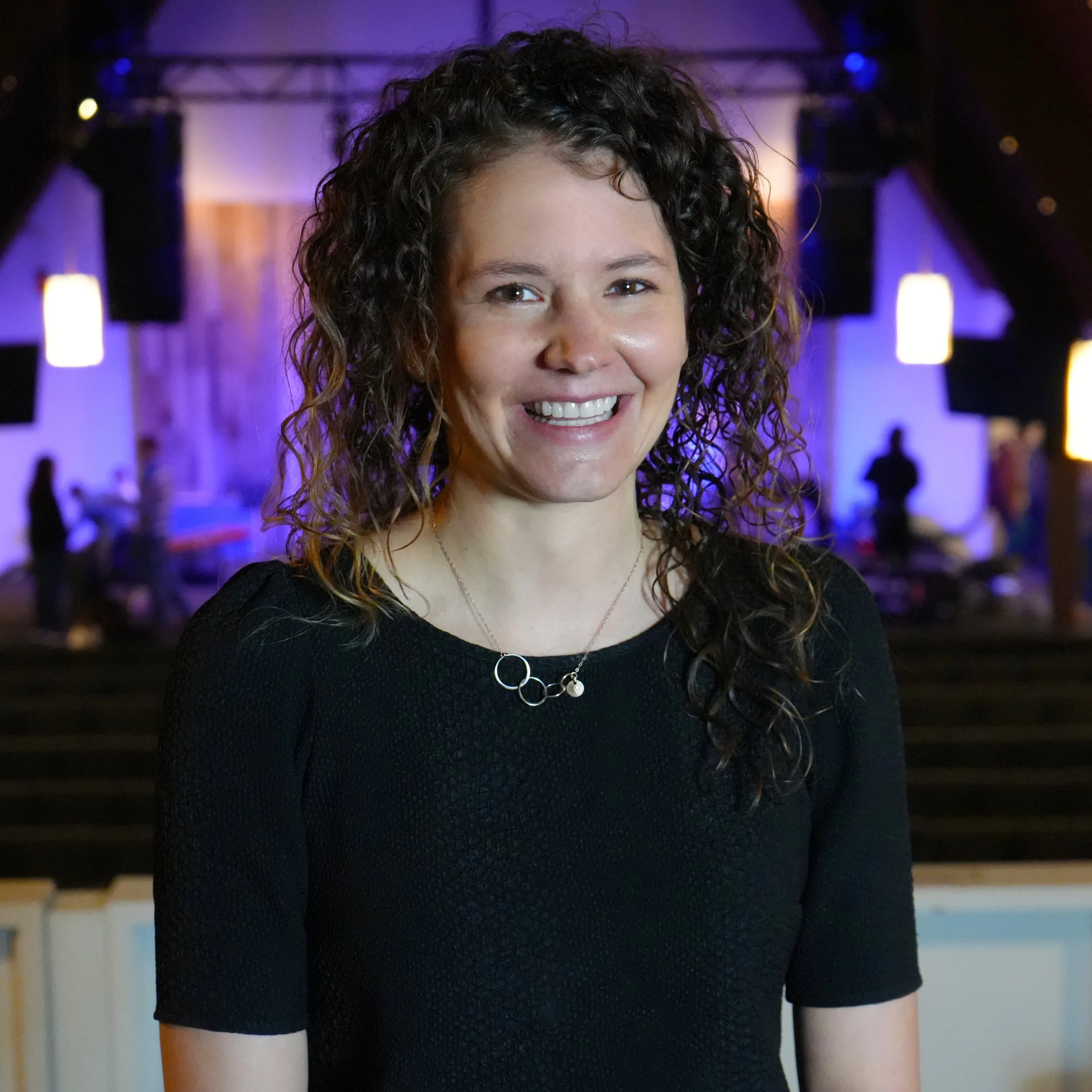 Smiling woman with curly hair stands in a dimly lit room, wearing a sleeveless purple dress.