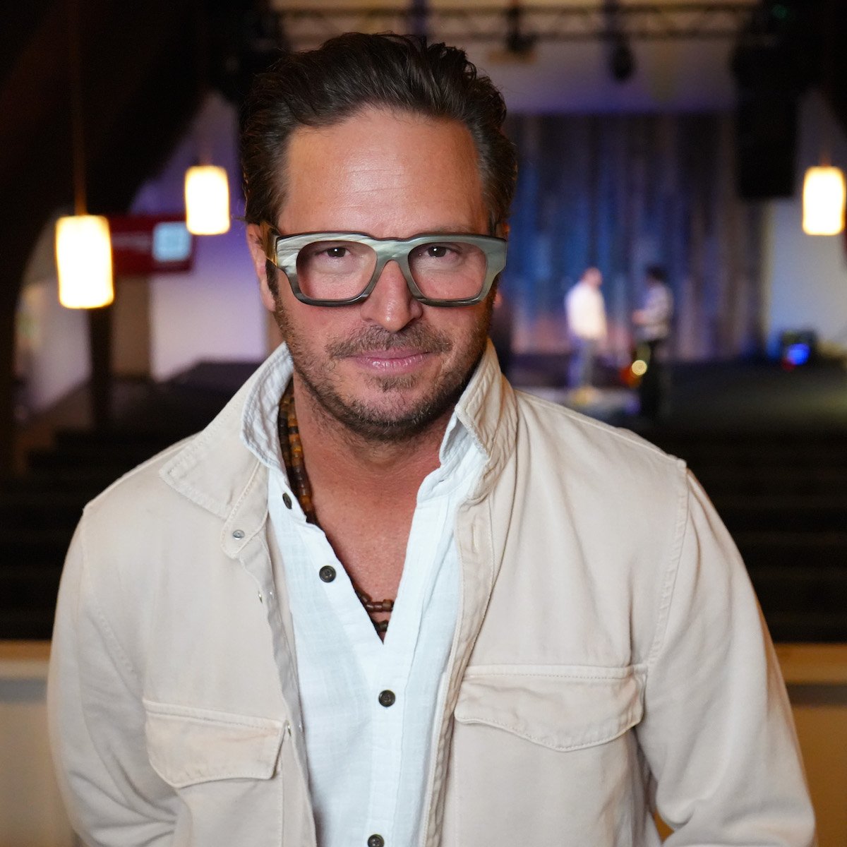 Man wearing glasses and a light jacket indoors with blurred background