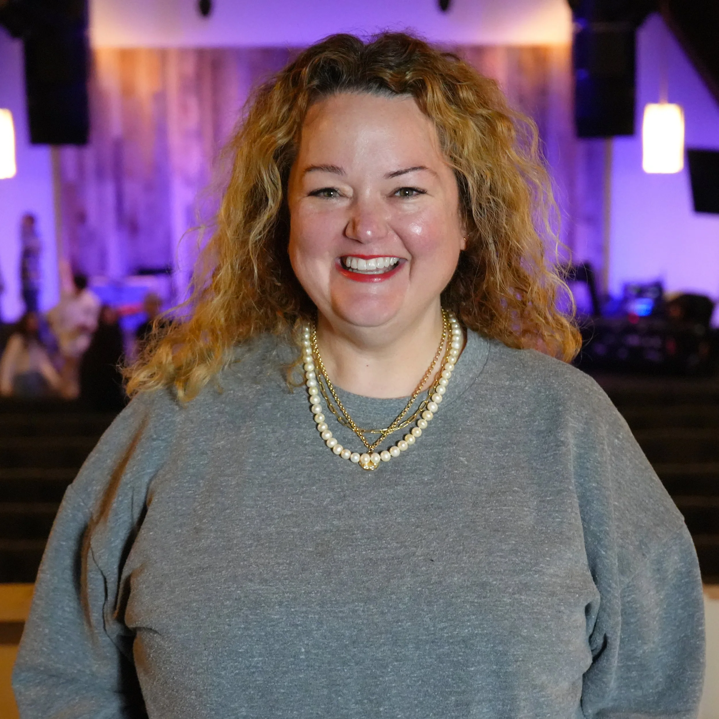 Woman with curly blonde hair wearing a purple cardigan in a dimly lit setting.