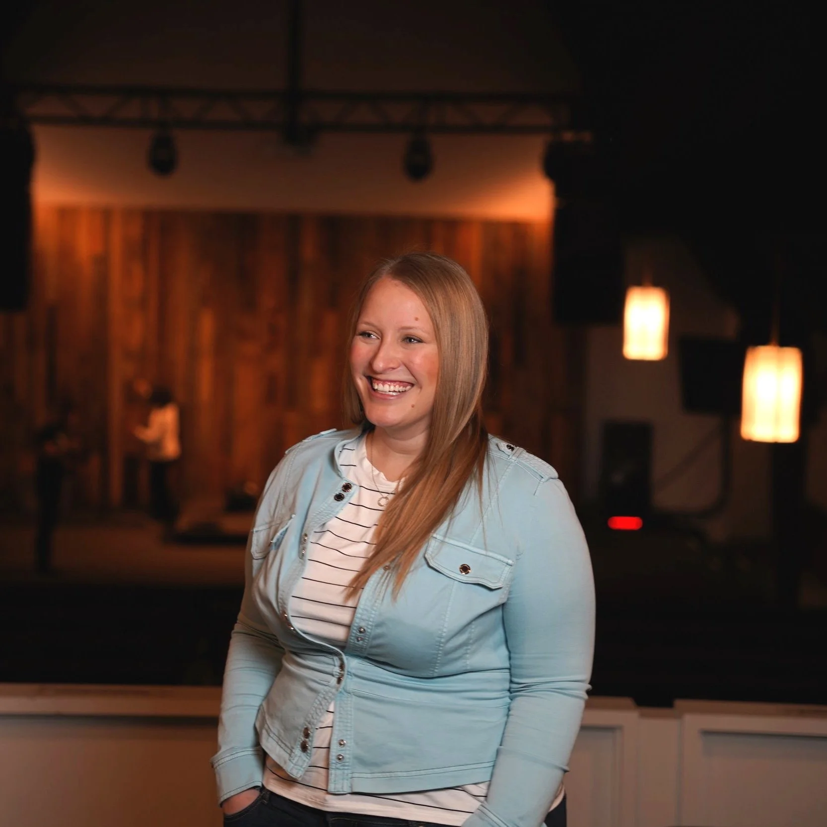 Smiling woman in a light blue jacket standing in a dimly lit room with blurred wooden background and hanging lights.