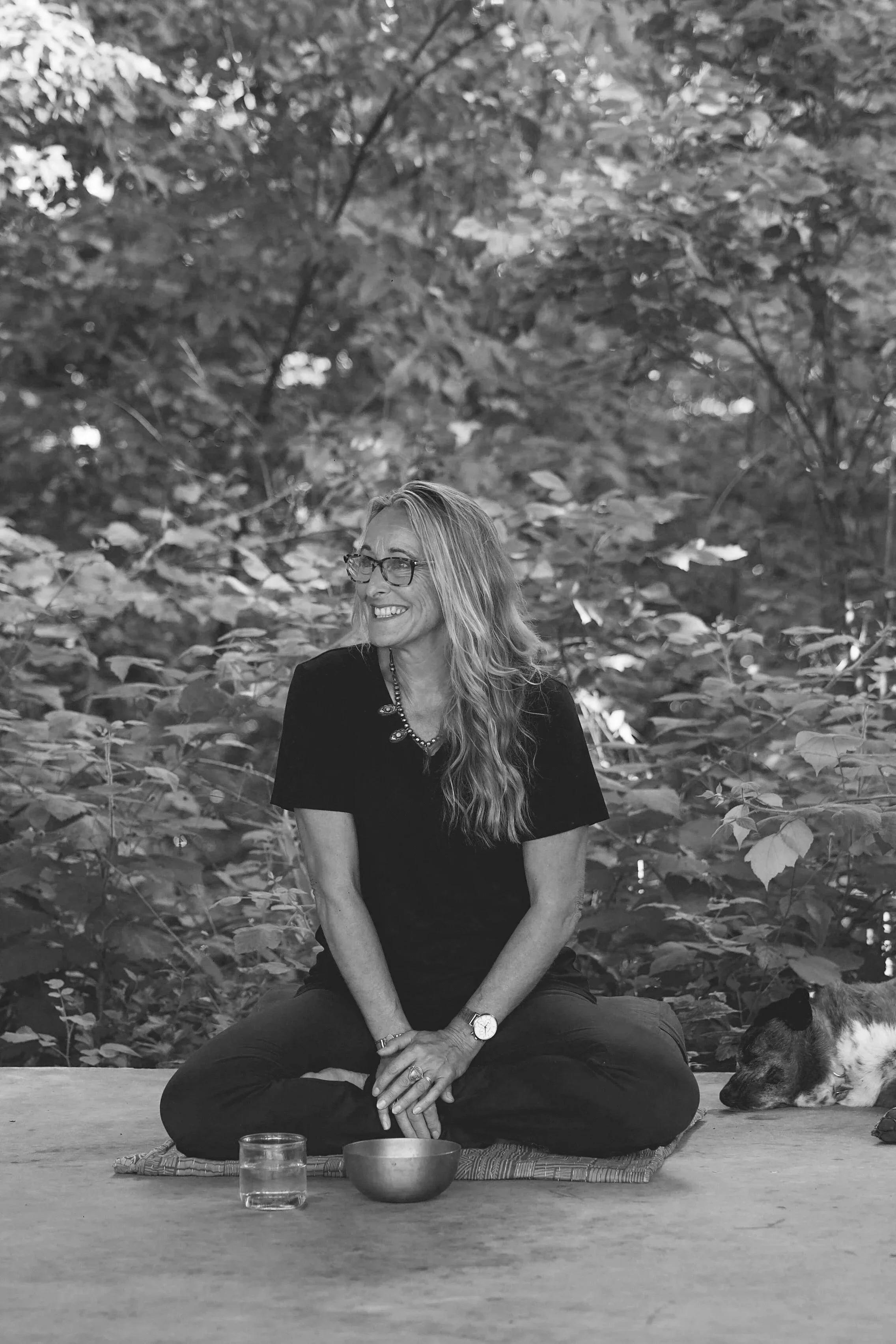 A woman with long hair, glasses, and a watch, sitting cross-legged on a mat in a forest setting, smiling. A dog is resting nearby, and there are bowls and a glass of water in front of her.