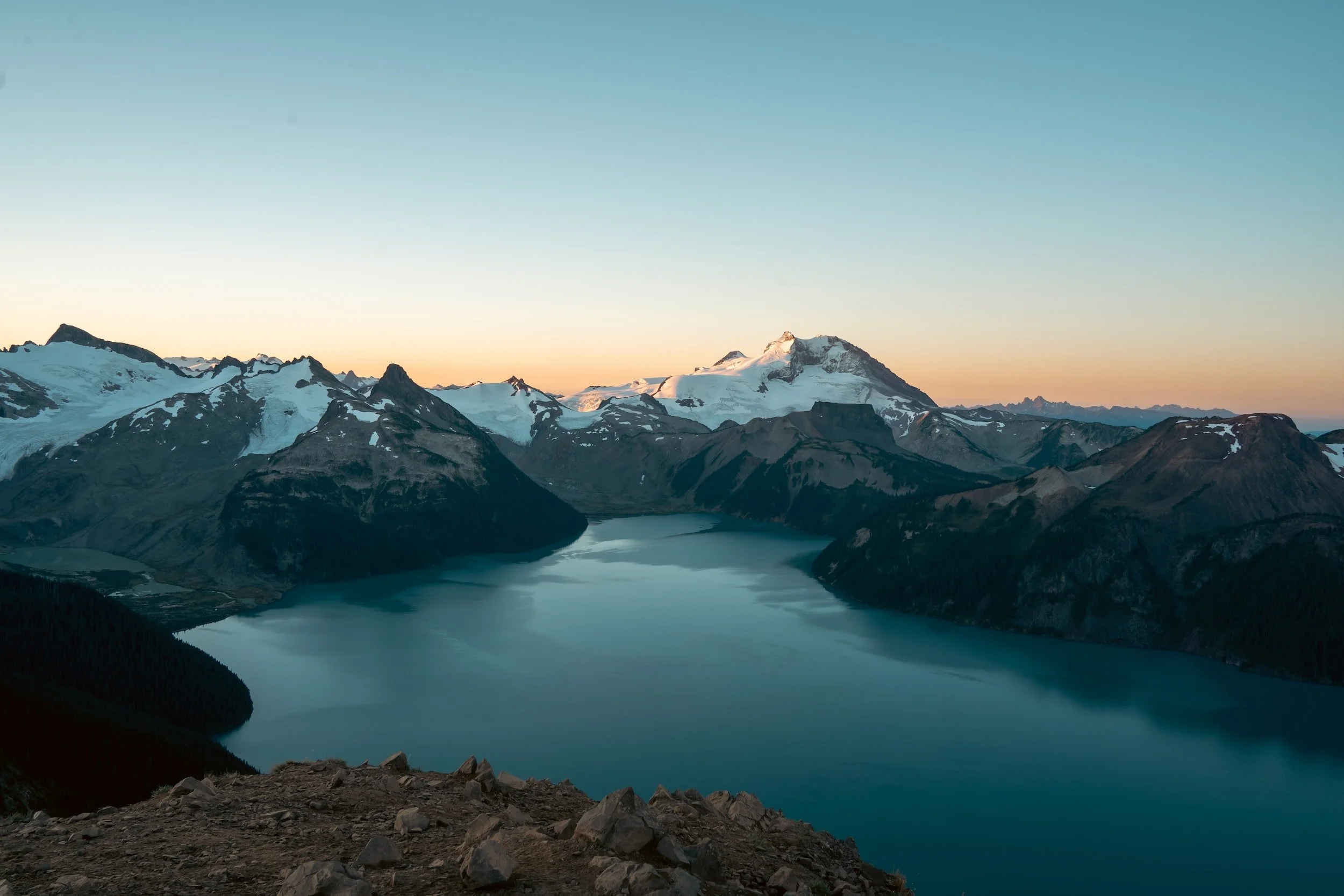 Scenic view of a serene lake surrounded by mountains with snow-capped peaks during sunset, under a clear sky.