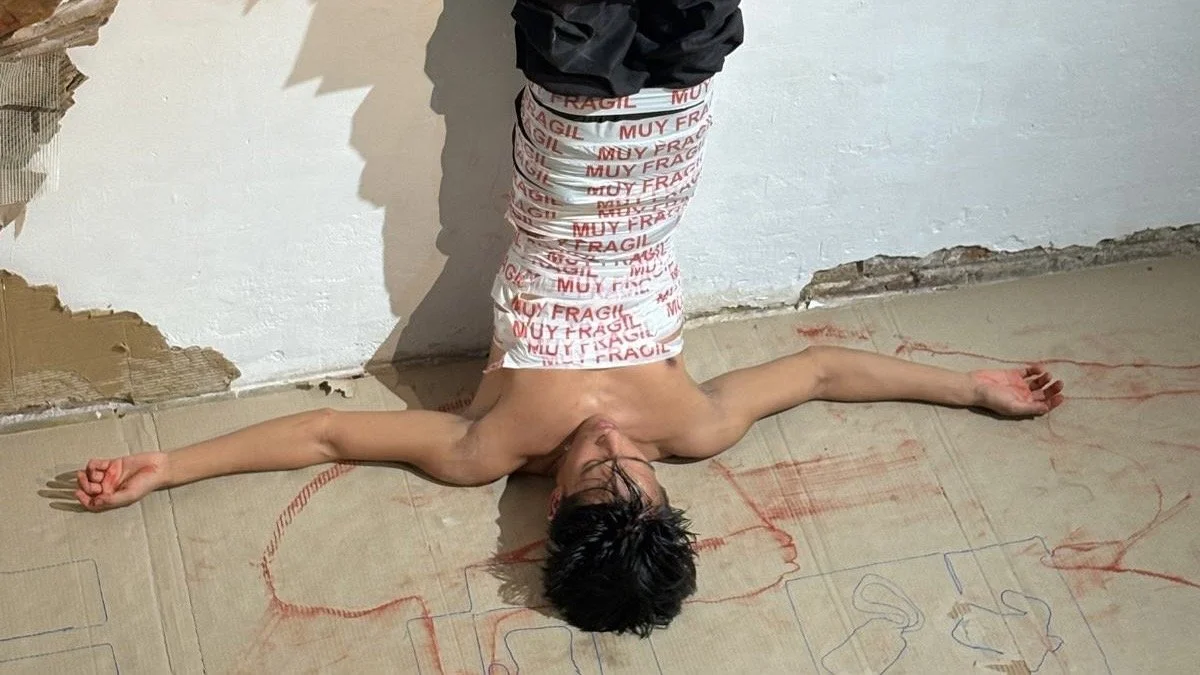 A shirtless person lying on their back on the floor with arms and legs spread, wearing white pants with red 'FRAGILE' and 'MUY FRAGILE' texts. The person's head is turned sideways, and they are on a surface with red and blue pencil drawings, next to a wall with damaged paint and plaster.