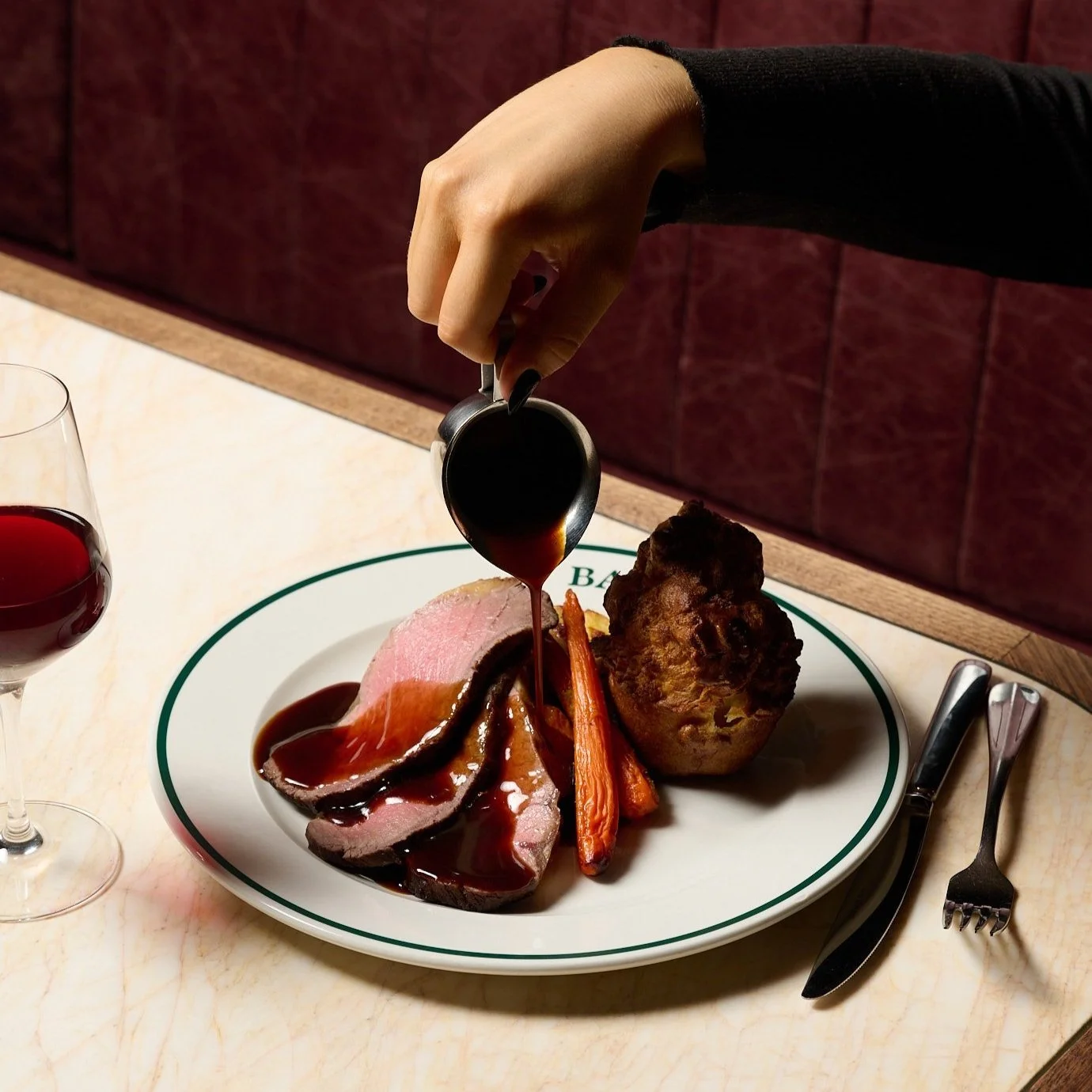 A person pouring red wine sauce over cooked meat and vegetables on a plate, with a glass of red wine and cutlery nearby.