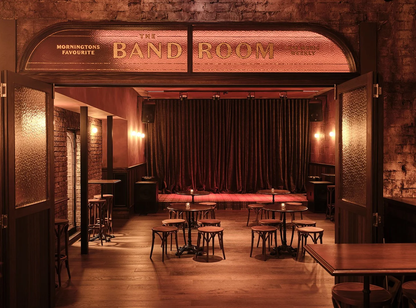 Interior of a cozy live music venue named "The Band Room," featuring a small stage with dark curtains, tables and chairs for audience, warm lighting, brick walls, and wood flooring.