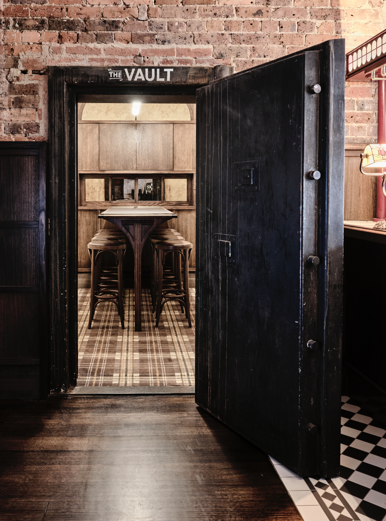 Interior view of a cozy cafe or restaurant with exposed brick walls, a dark wood door marked "The Vault," and a small wooden table with stools inside a private booth or room.