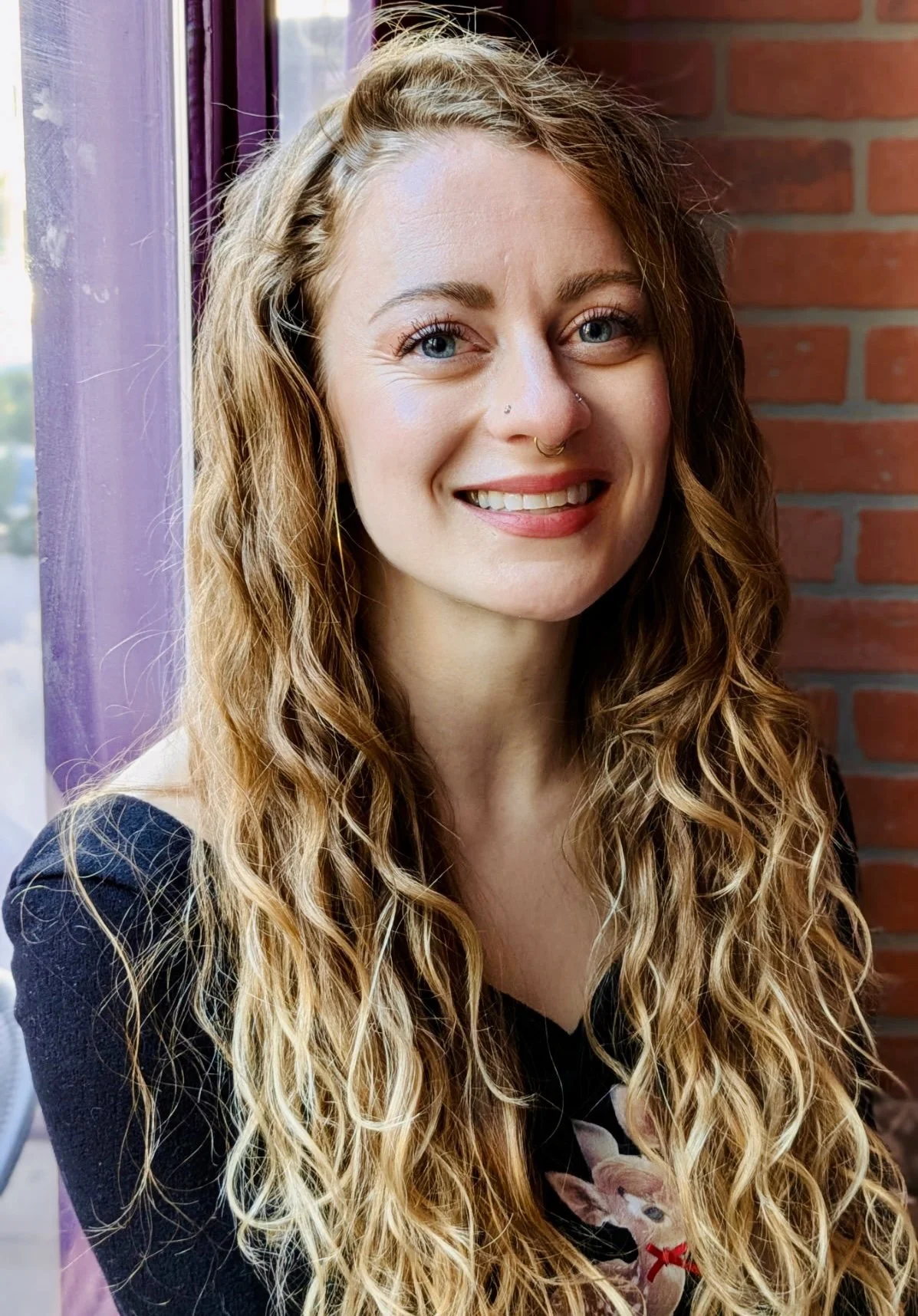 A woman with long, curly blonde hair and blue eyes, smiling, standing near a purple window frame and a brick wall, wearing a black top.