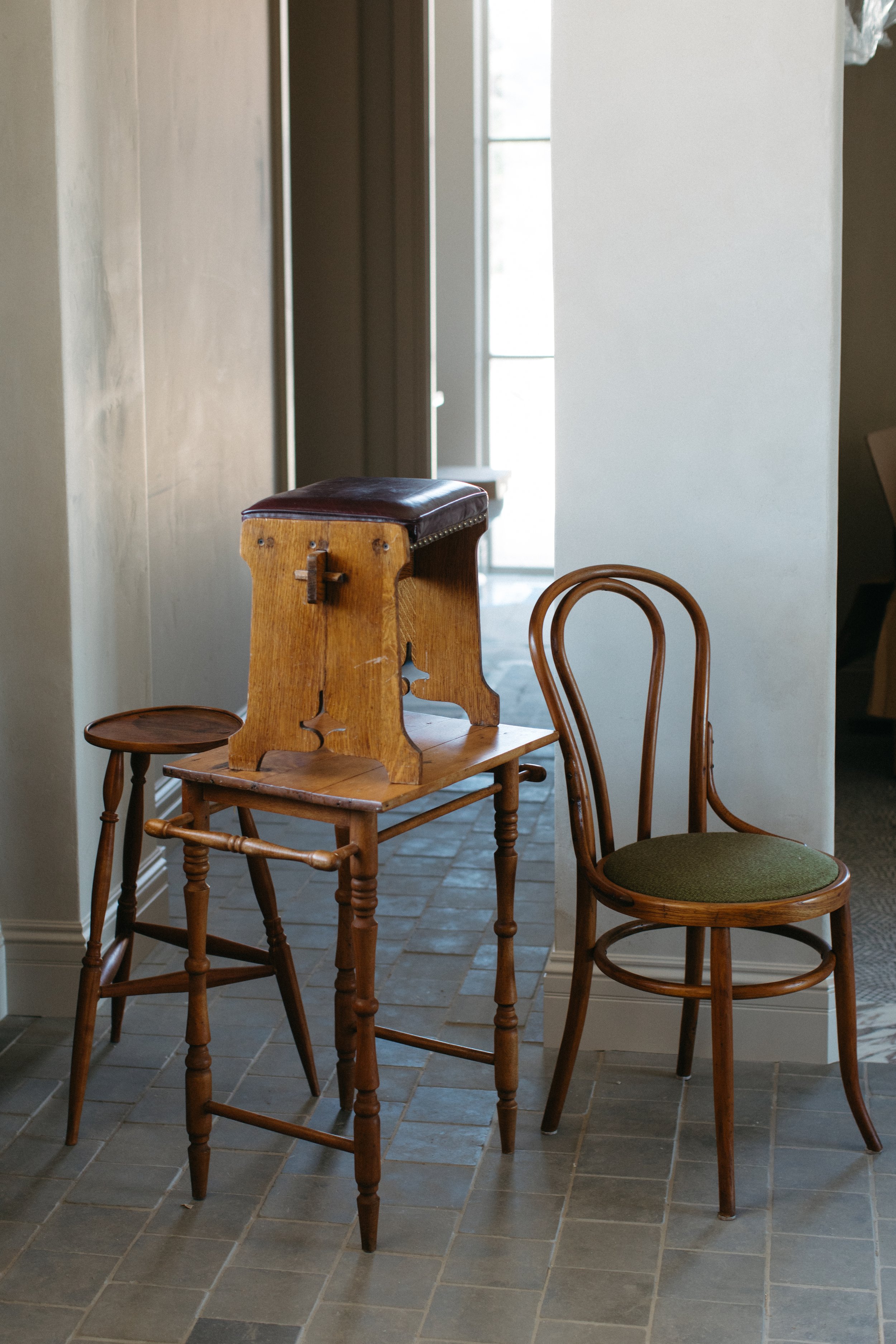 Two wooden chairs, one with a padded cushion on top, are placed against a white wall. The chairs are of different styles and sizes, with a background of a door and stone-tiled floor.