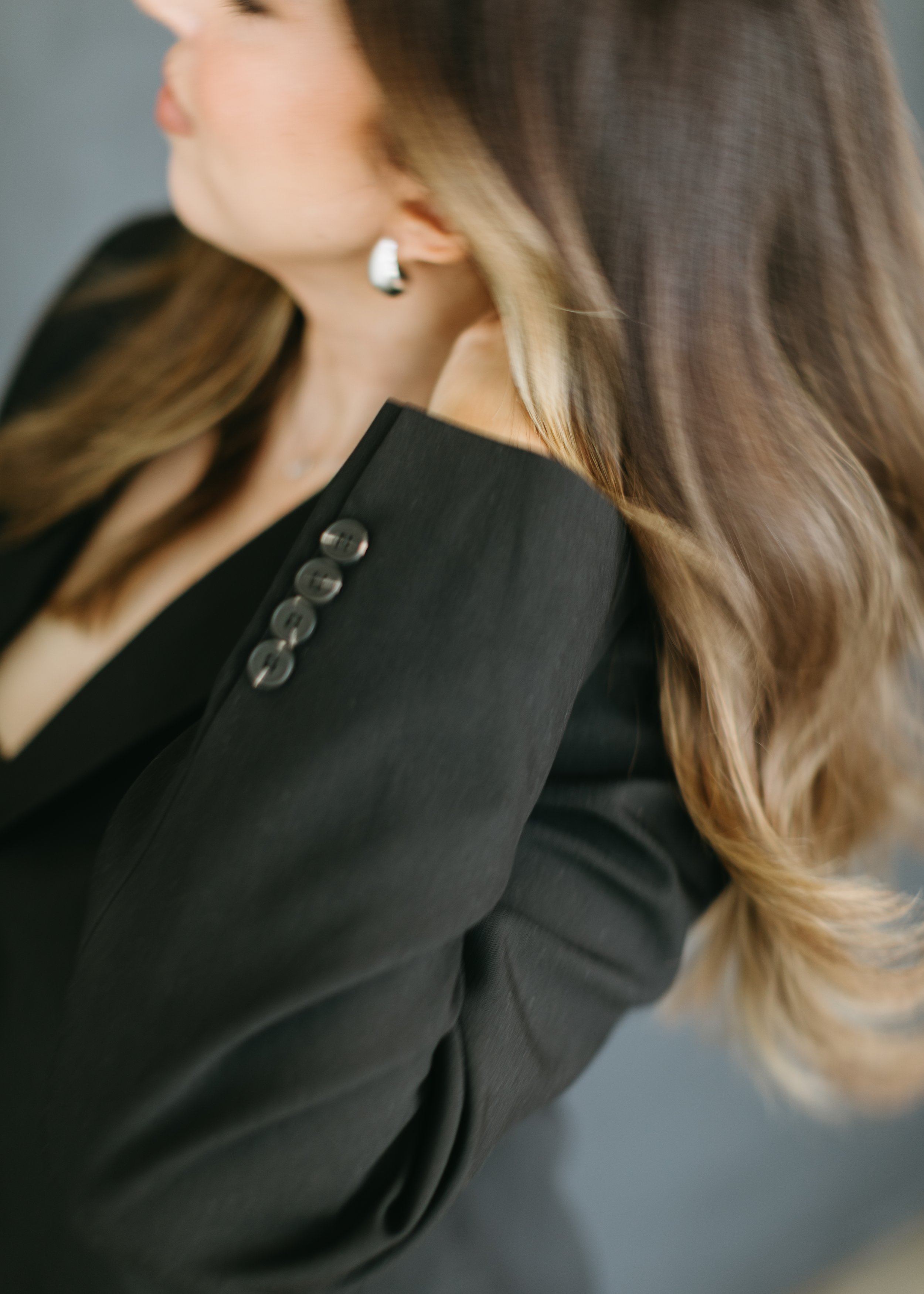 Close-up of a woman wearing a black blazer with button details on the sleeve, with long, wavy, brown hair and silver hoop earrings, touching her neck.