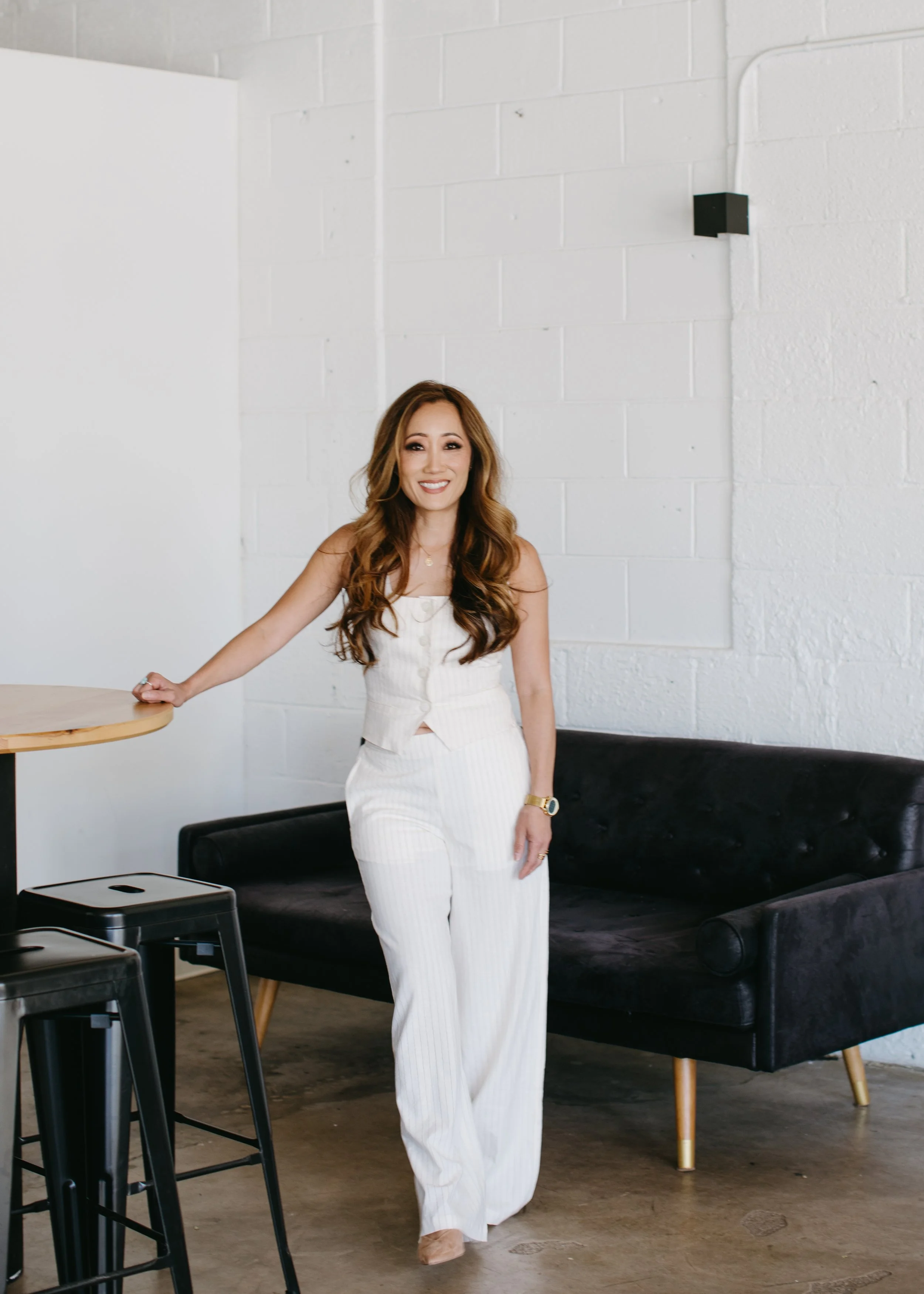 A woman with long, wavy brown hair standing in a modern, minimalistic room with white brick walls. She is smiling and wearing a white sleeveless top and matching wide-leg pants, accessorized with a watch and jewelry. She is leaning on a small round w