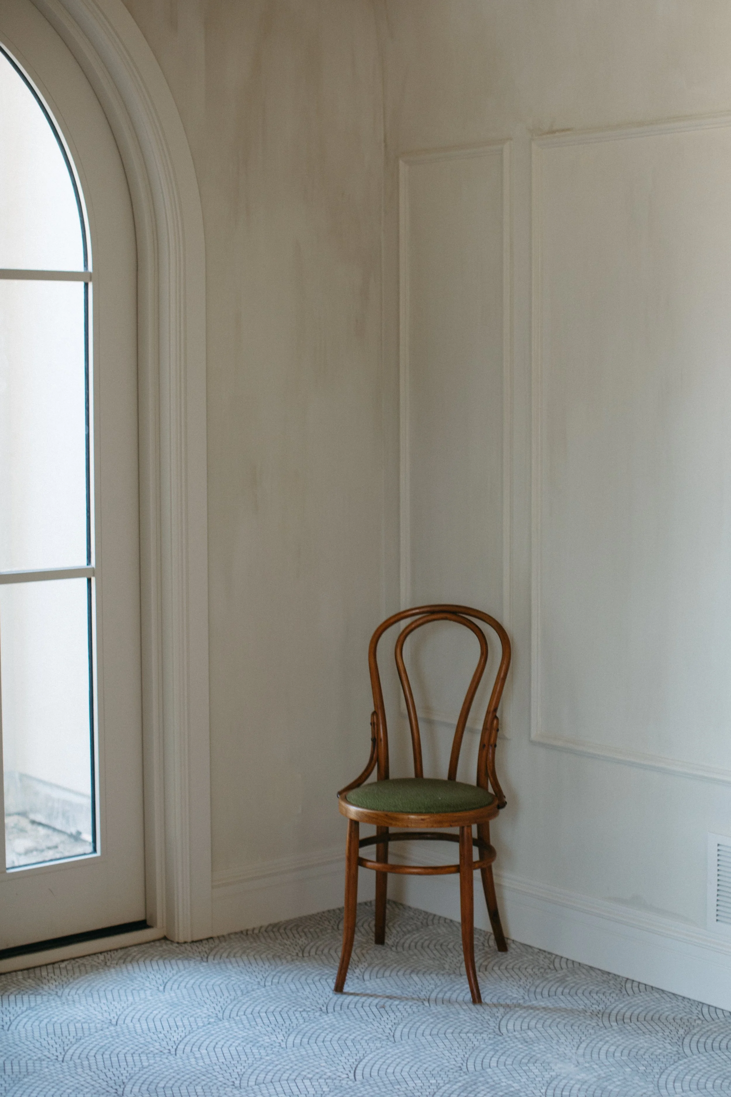A vintage wooden chair with a green upholstered seat in a corner next to a window and plain white walls.