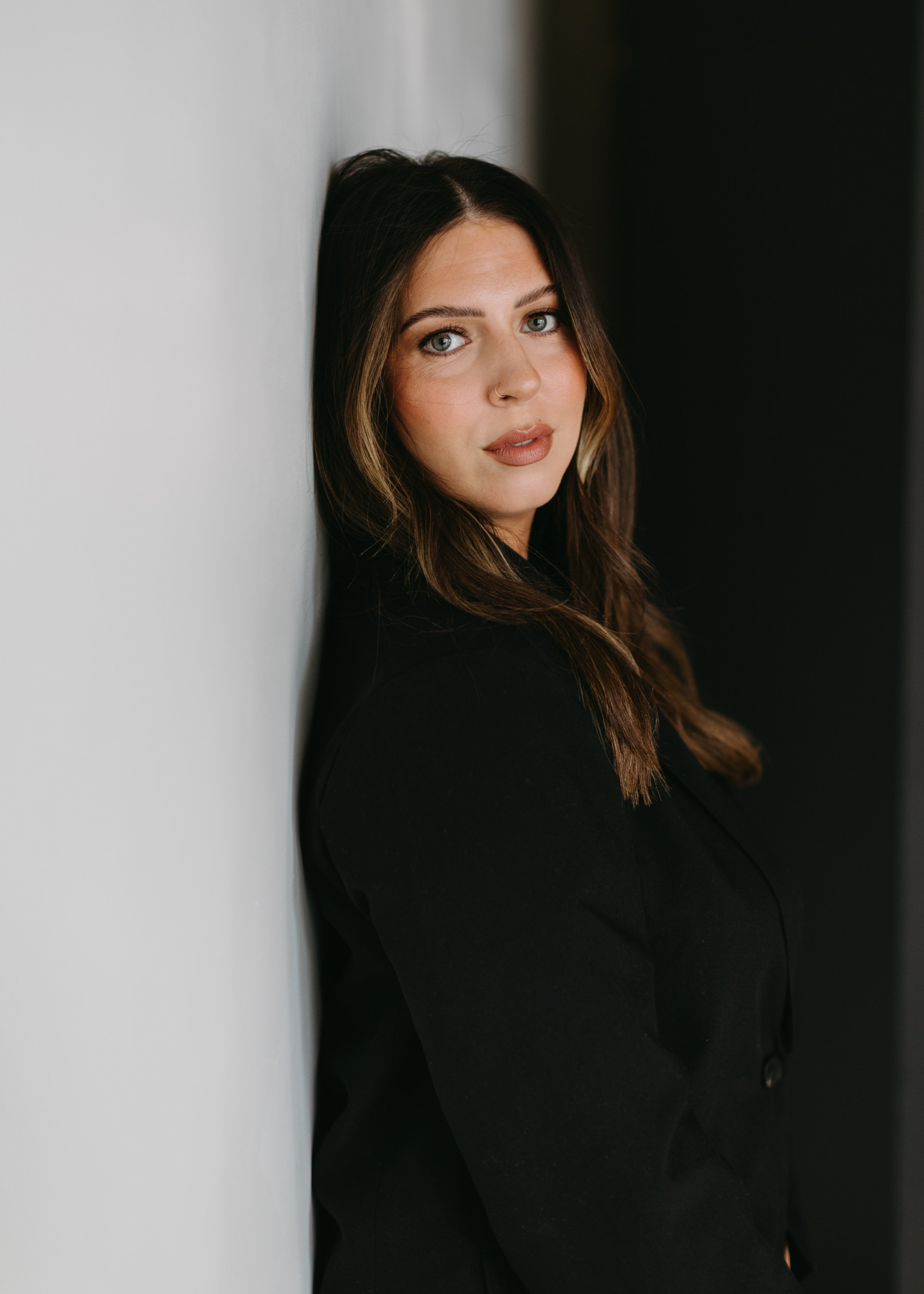 A woman with long brown hair, wearing a black top, leaning against a wall with a neutral expression.