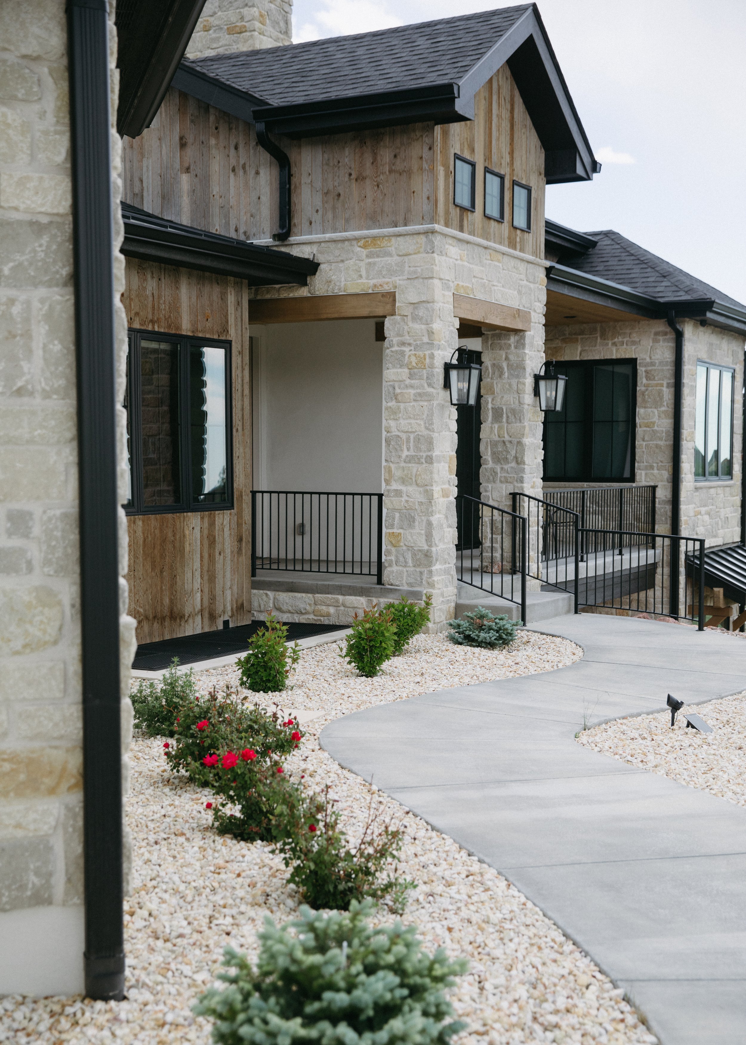 Front view of a modern house with stone and wood exterior, black railings, a sidewalk, and landscaped garden with small bushes and flowers.
