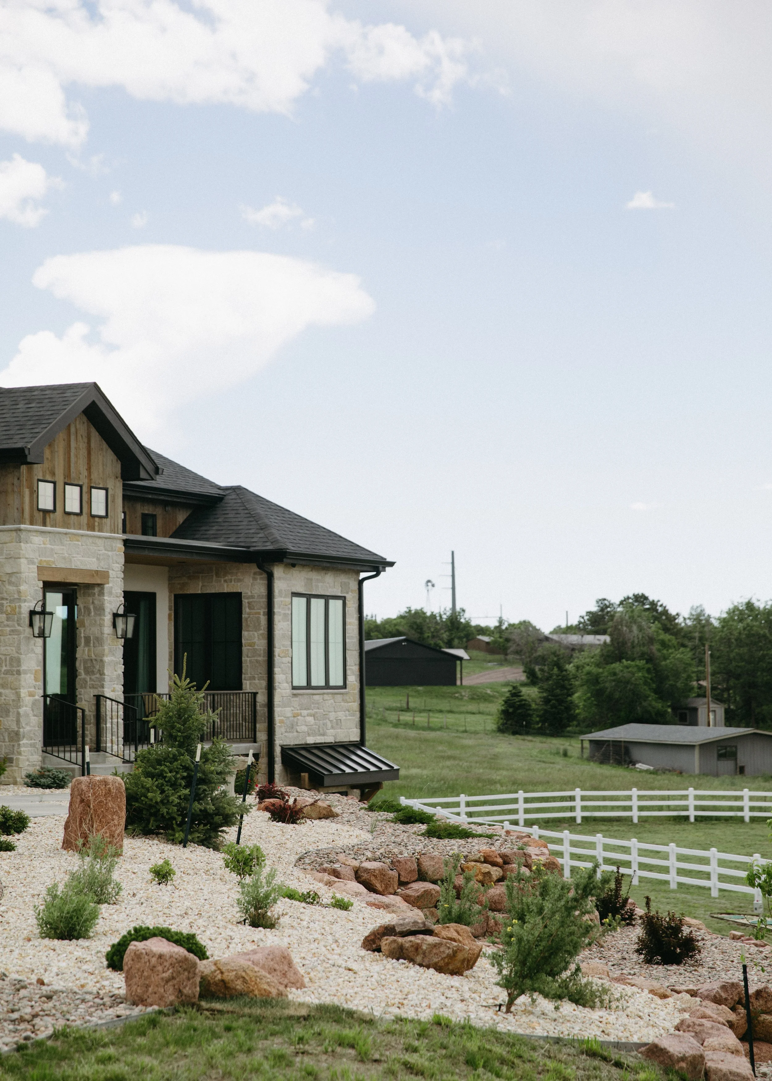 Front yard landscape with rocks, small shrubs, and young trees in front of a modern house with stone and wood exterior, black window frames, and black roof, under a partly cloudy sky.