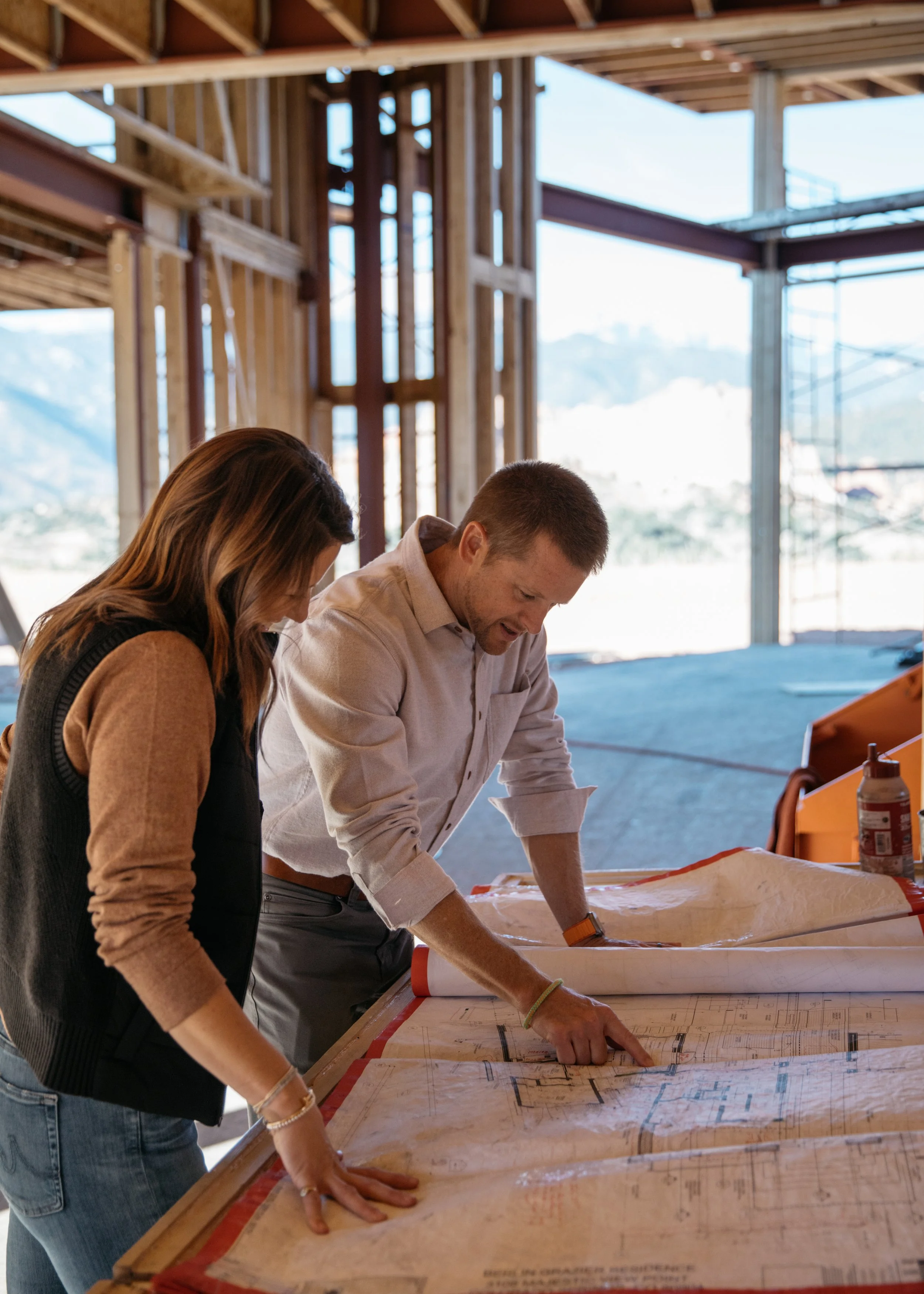 Two people examine blueprints on a construction site with wooden framing and large windows, mountain scenery in the background.