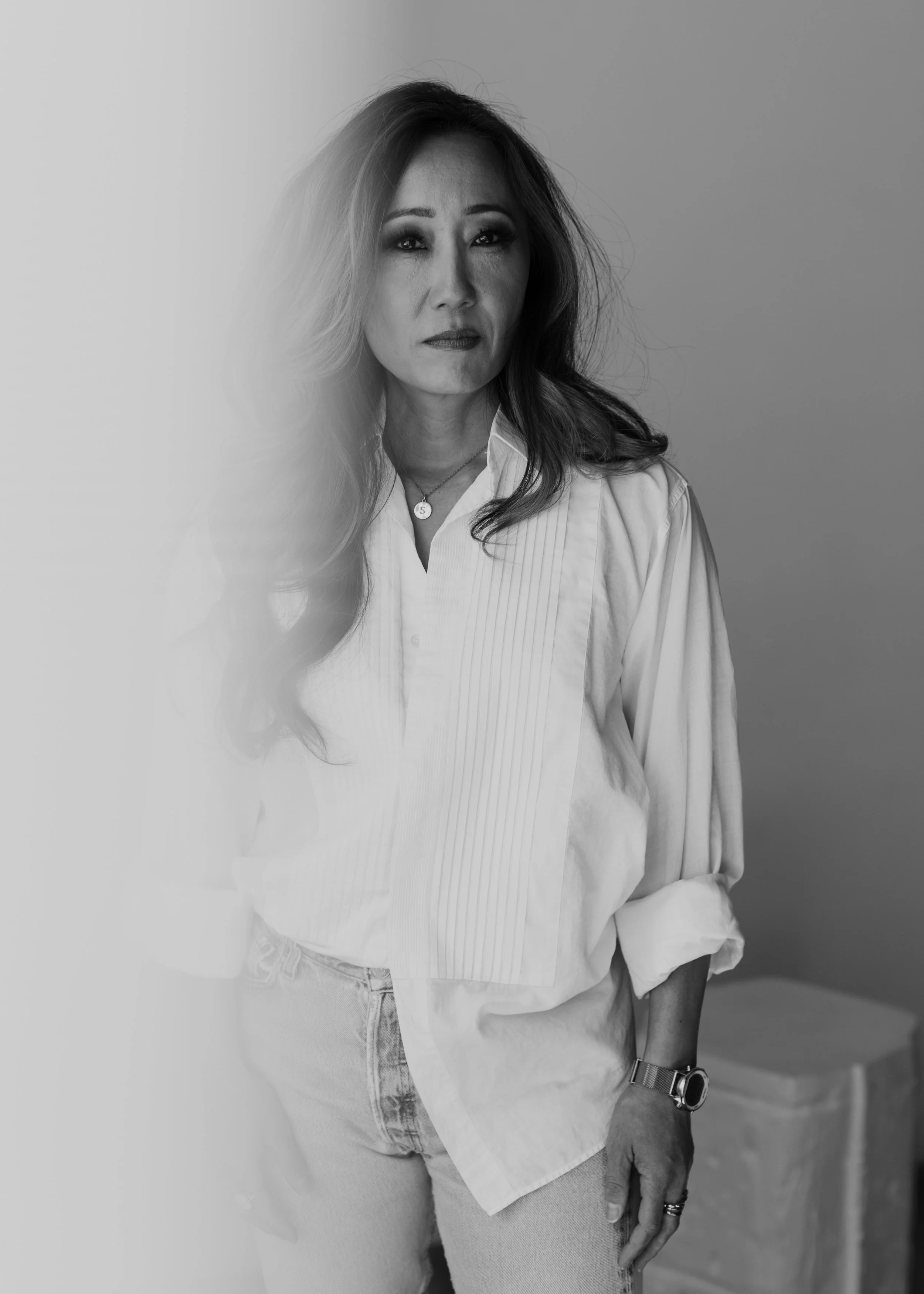 A black and white photo of a woman with long wavy hair, wearing a white button-down shirt, jeans, a watch, and a necklace, standing with one hand in her pocket against a plain background.