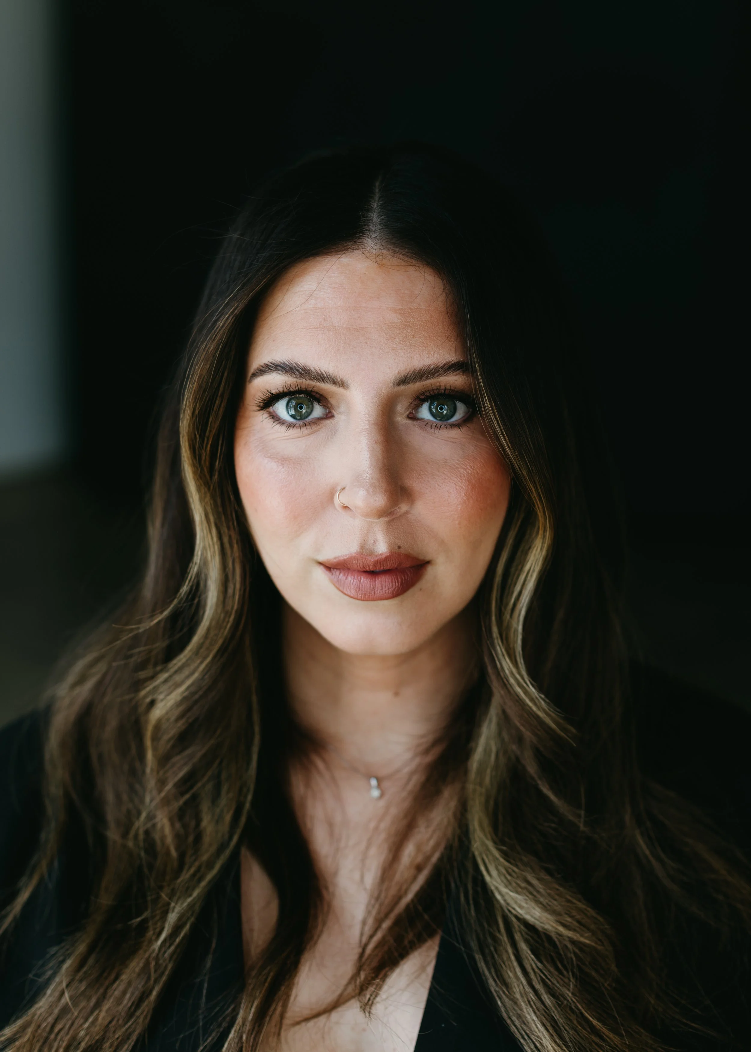 Close-up of a woman with long wavy brown hair and blue eyes, wearing makeup and a small nose ring, against a dark background.