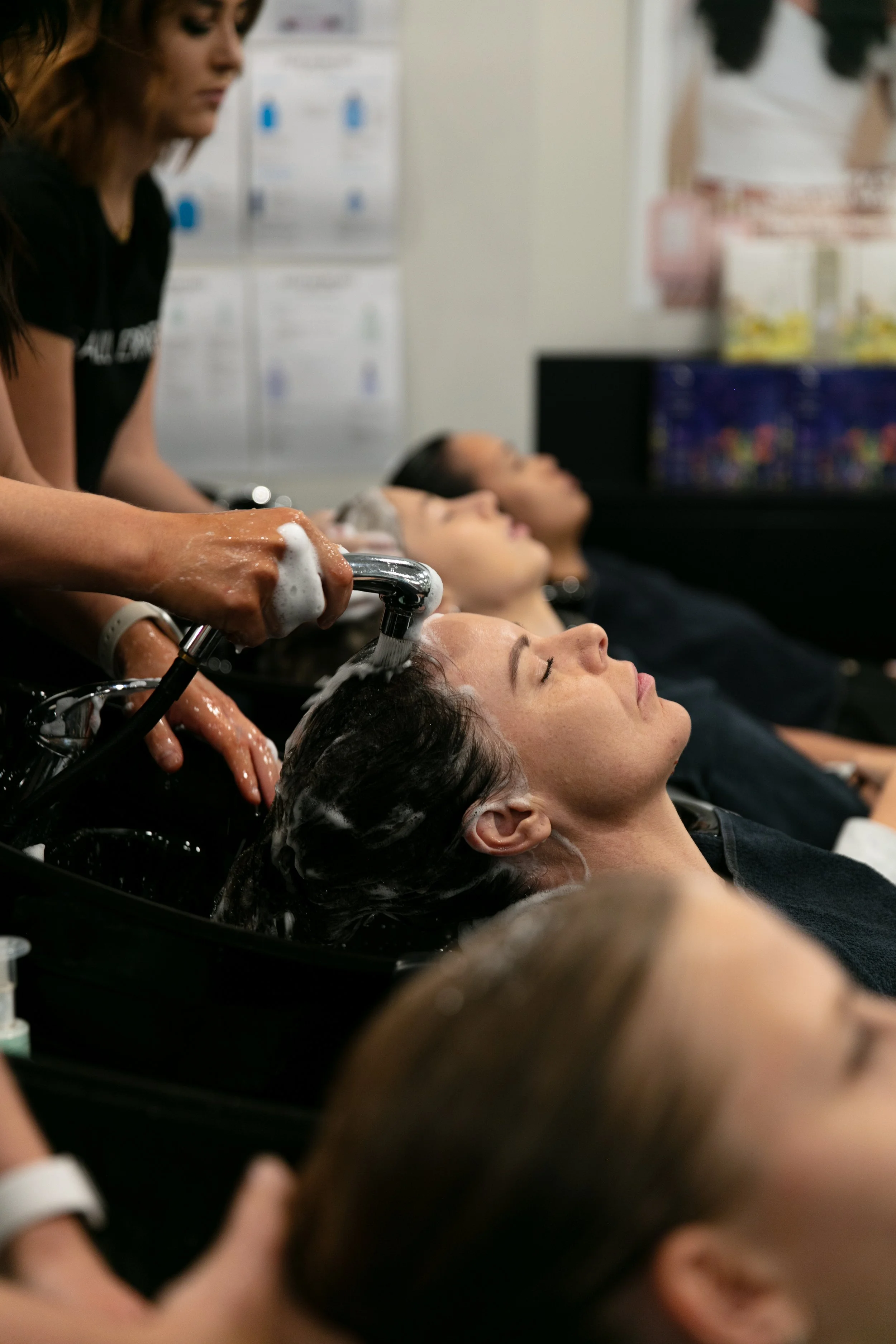 People reclining with their heads in salon wash basins while someone shampoos their hair.